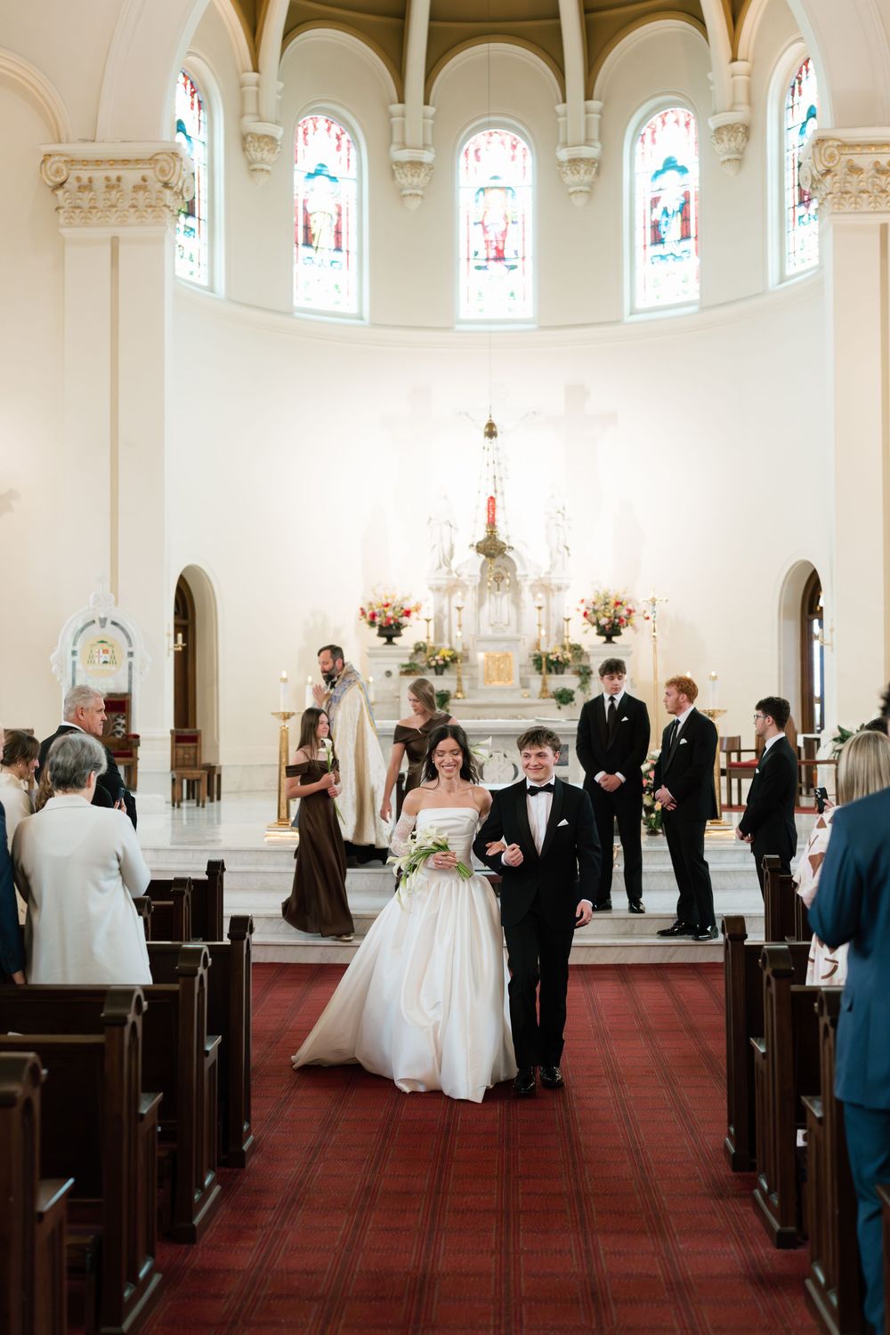 Bride and groom walk down the aisle of a white church with stained glass windows after their wedding ceremony.