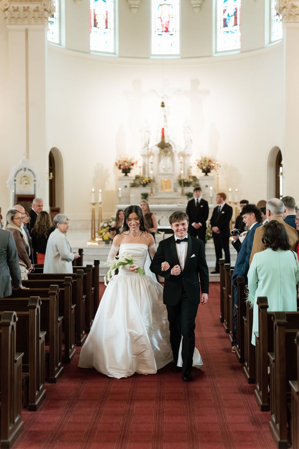 Bride and groom walk joyfully down church aisle after wedding ceremony, guests standing in pews, red carpet, ornate altar.