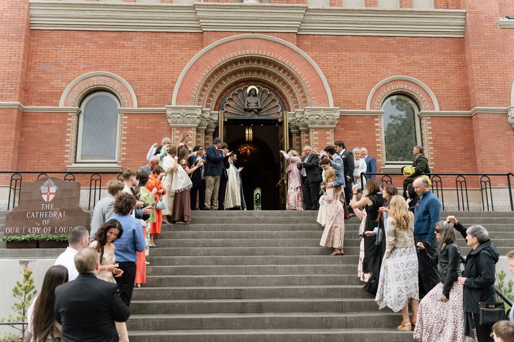 Wedding couple exits brick church greeted by cheering guests on stone steps during outdoor ceremony celebration.
