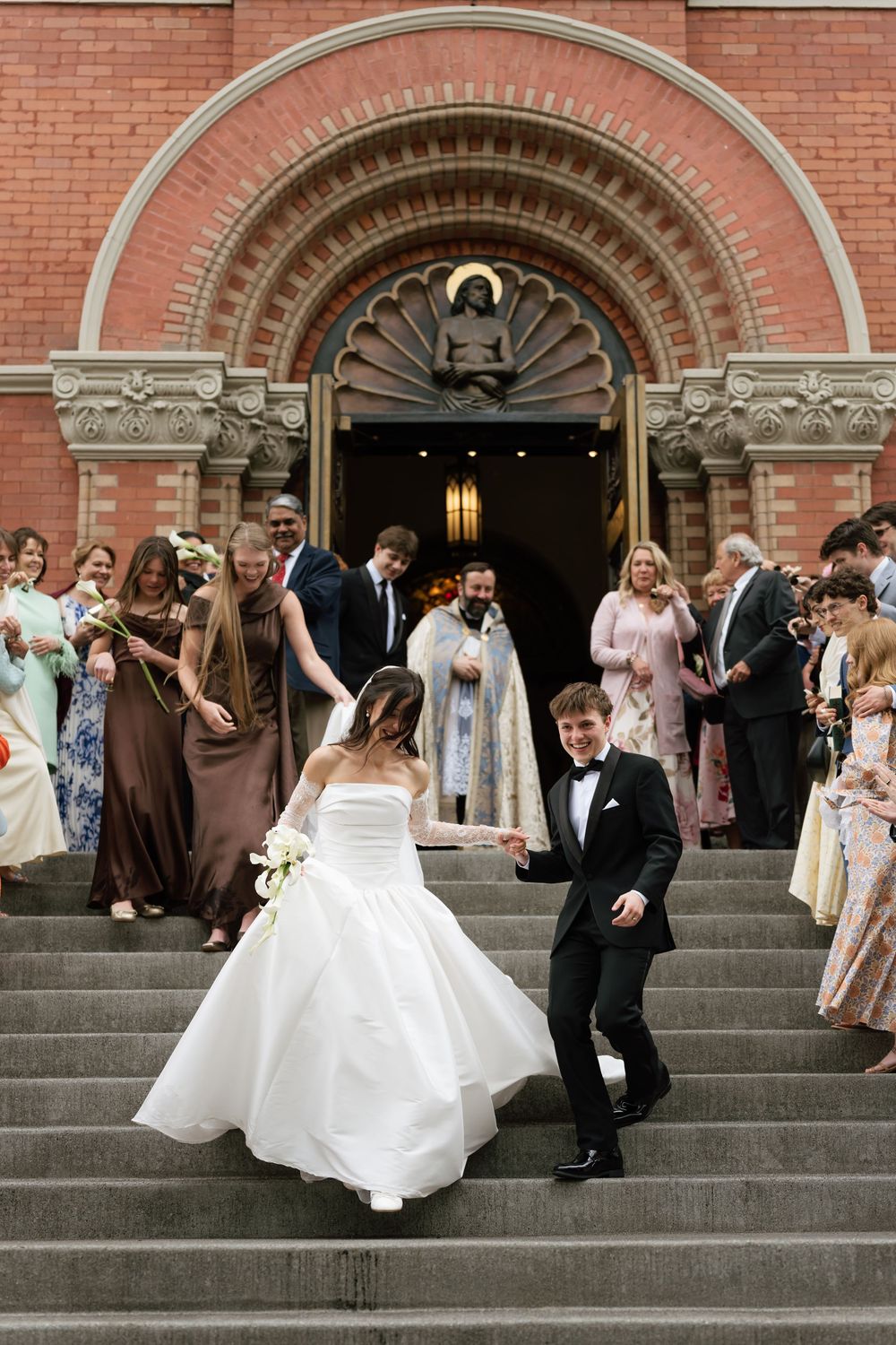 Bride and groom descend church steps after wedding ceremony, surrounded by guests and officiant at red brick church entrance.
