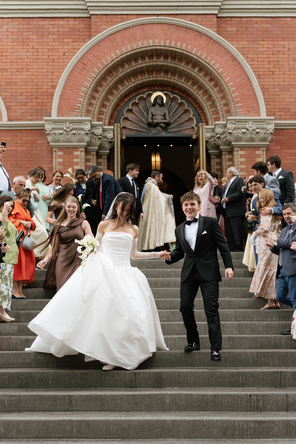 Newlyweds descend church steps hand-in-hand as guests celebrate outside a red brick church after their wedding ceremony.