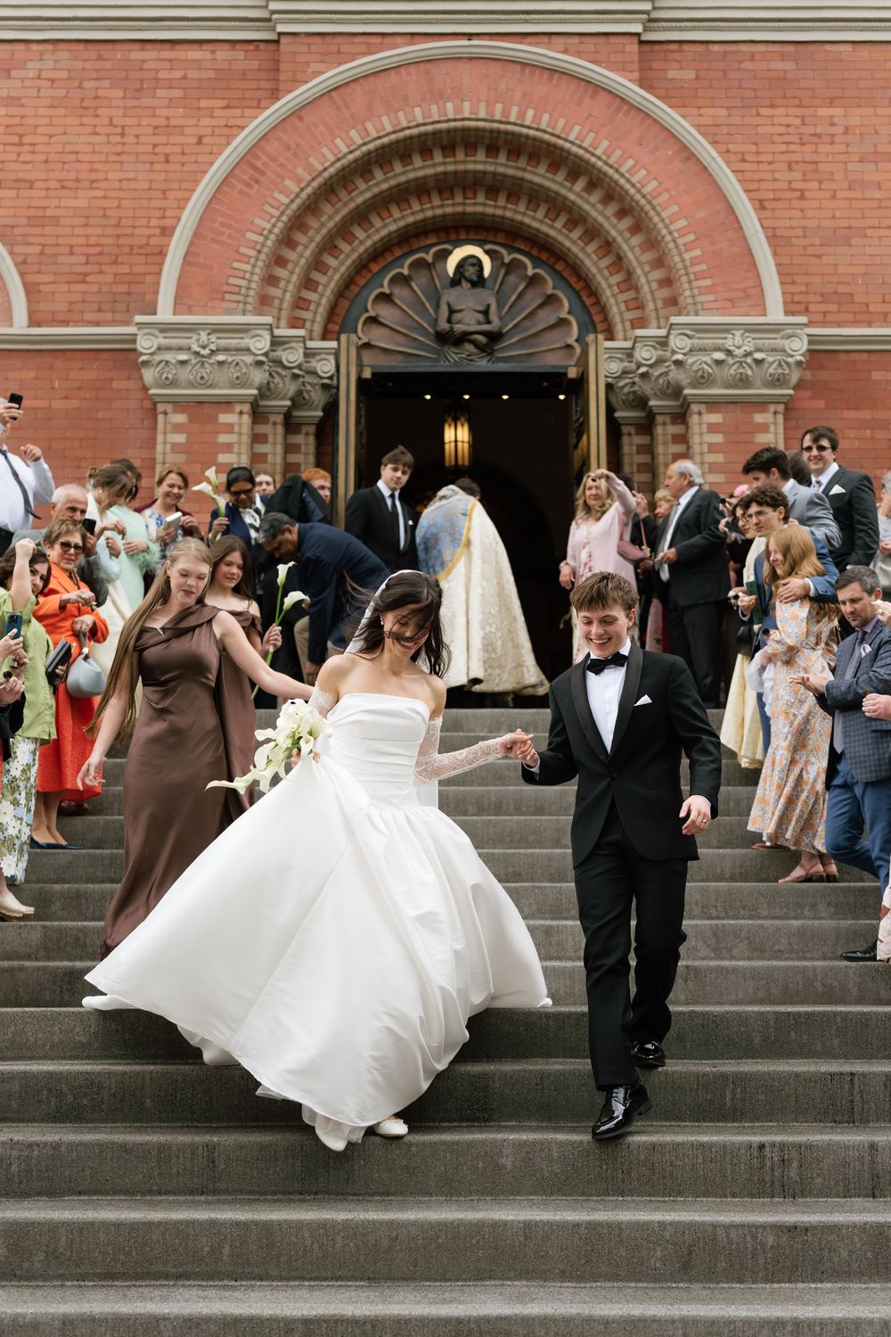 Bride and groom descend church steps in wedding attire, guests celebrating behind them at a red brick church.