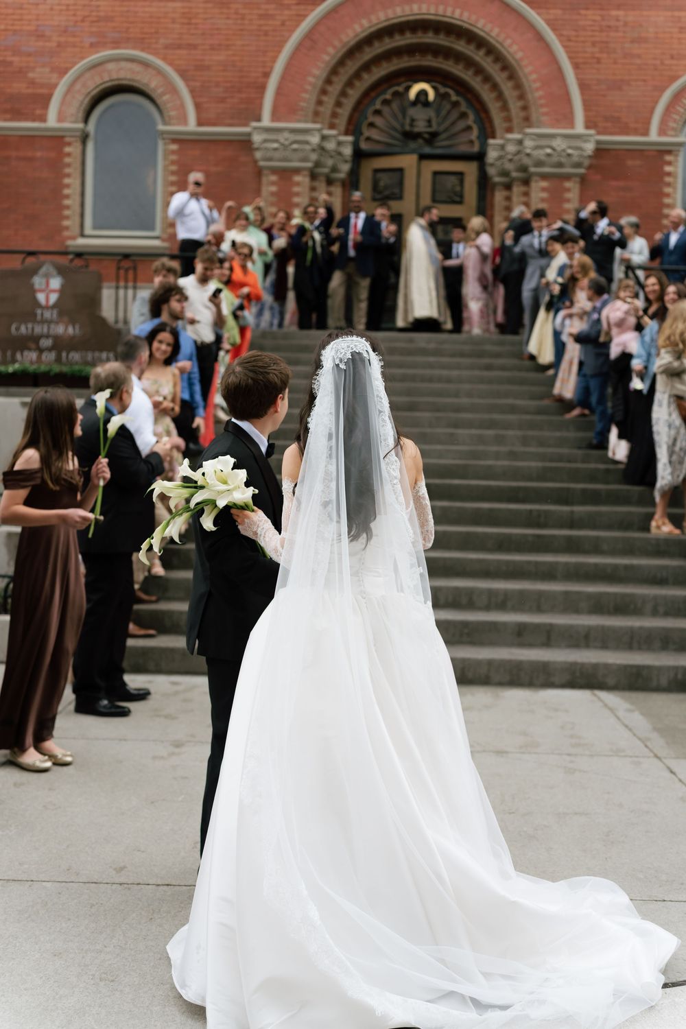 Bride in white gown with veil and groom in tuxedo exit a historic cathedral surrounded by wedding guests.