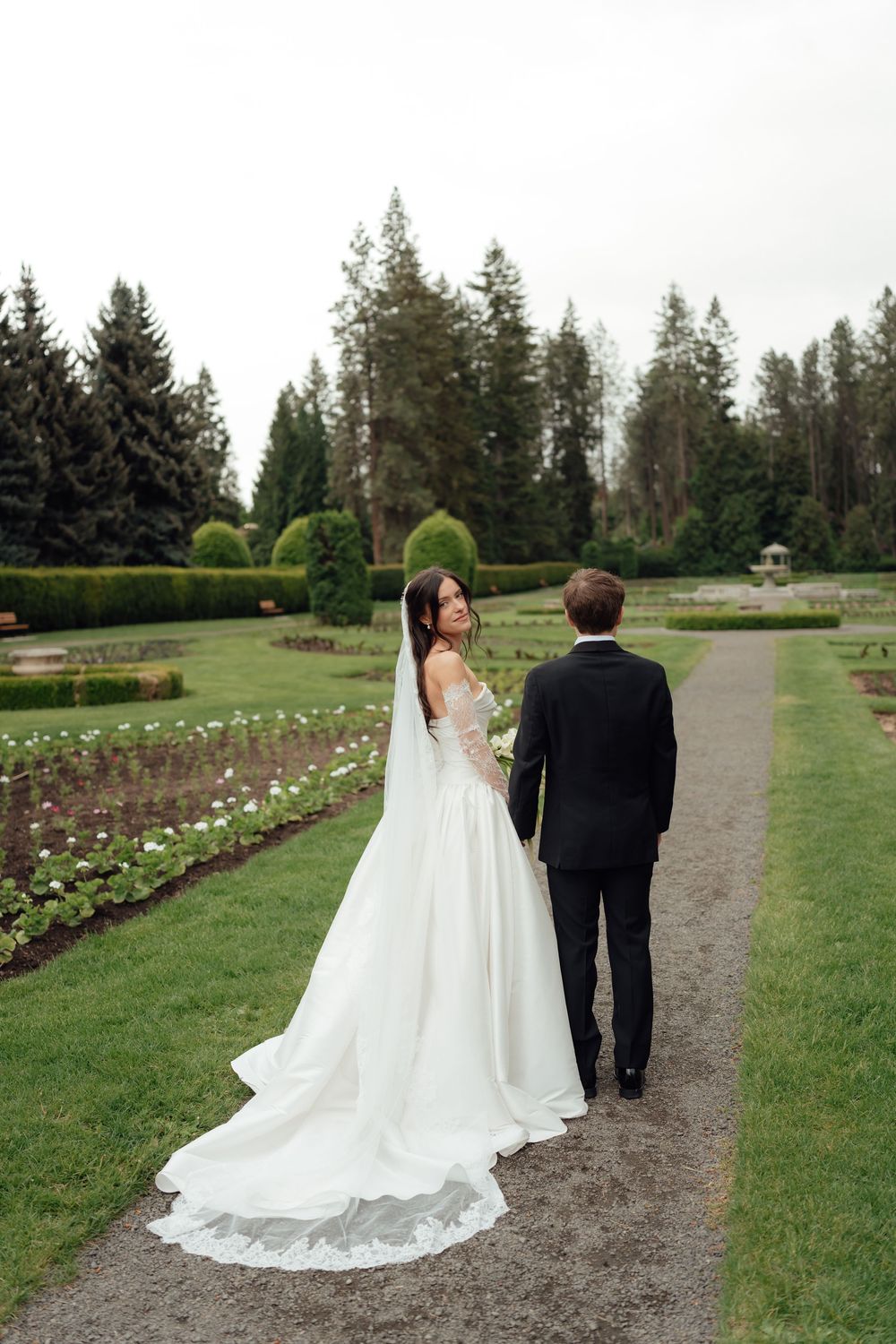 Bride in white gown with veil and groom in black suit walk hand in hand along garden path surrounded by lush greenery.
