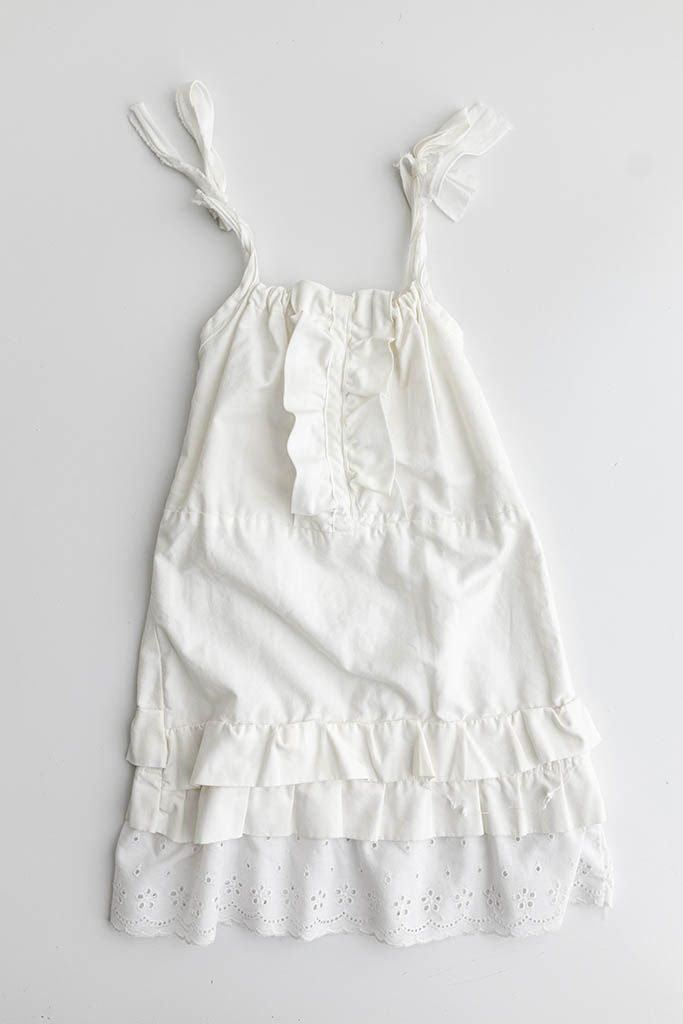 White cotton sundress with tie straps, ruffle front detail, tiered skirt and eyelet lace hem, laid flat.