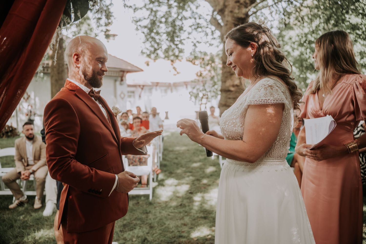 Outdoor wedding ceremony with groom in burgundy suit exchanging vows with bride in white dress beside officiant.