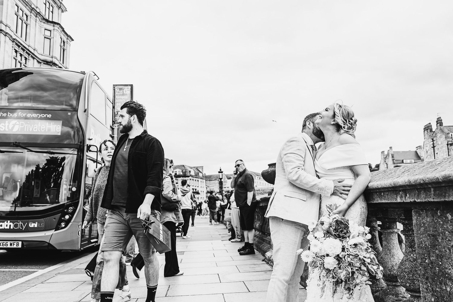 Black and white photo of a couple kissing on a busy city street, bride holding flowers, bus and pedestrians in background.