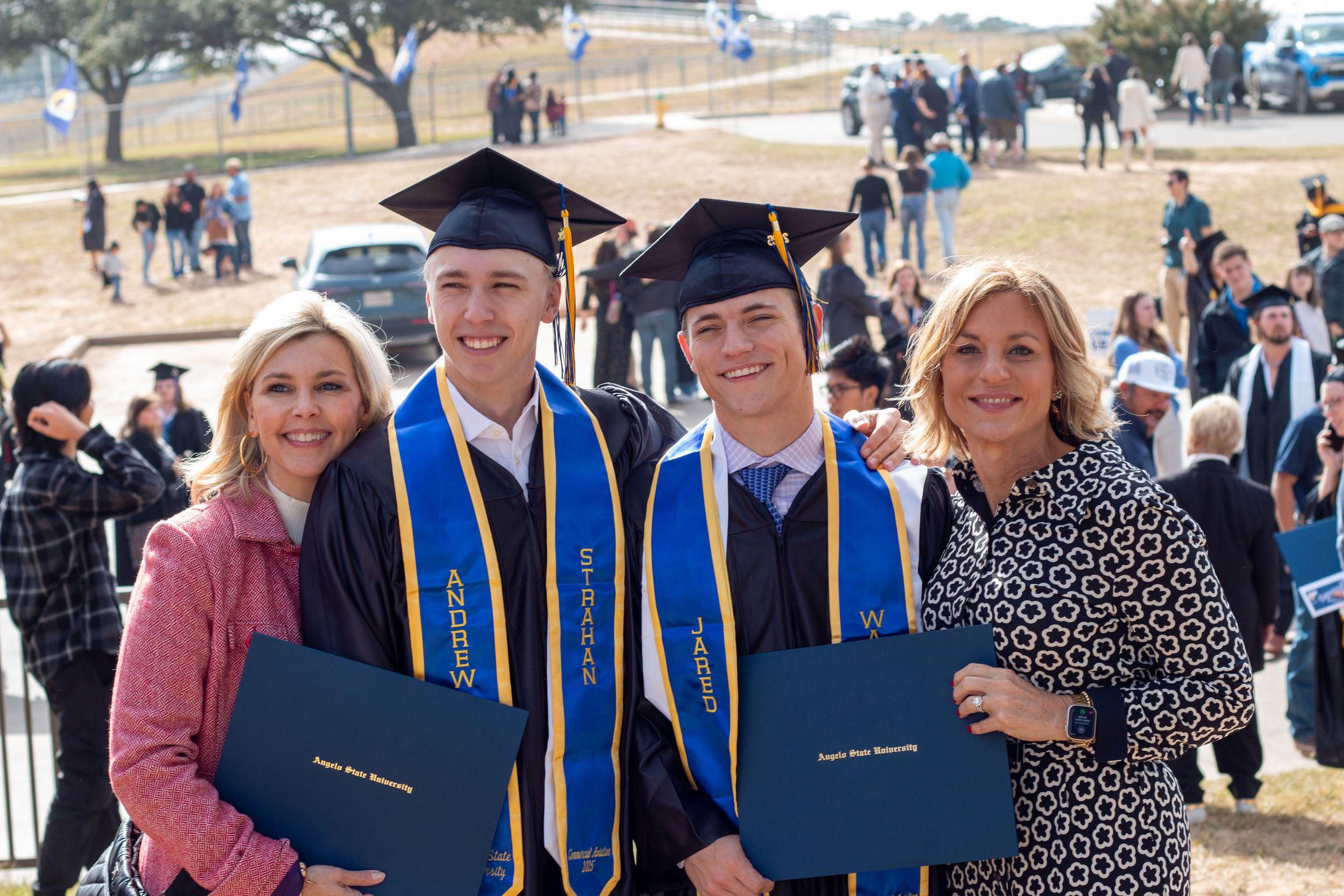 Two graduates in cap and gown hold diplomas with proud family members at an outdoor college graduation ceremony.