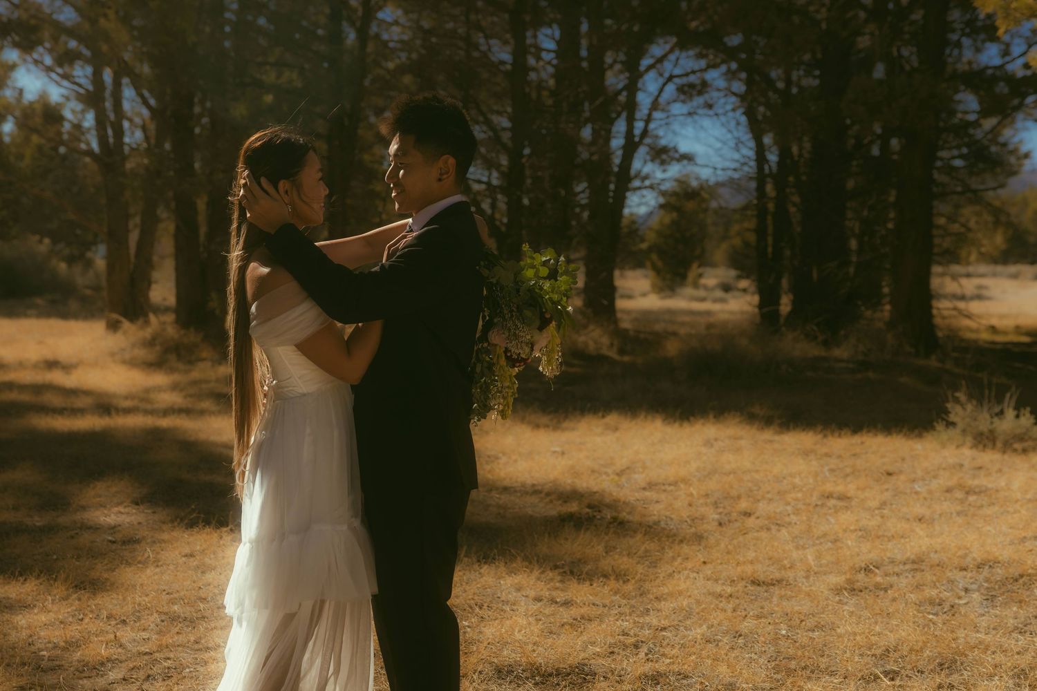 A bride and groom share a romantic embrace in a sunlit forest meadow, the bride in a white gown holding a bouquet.