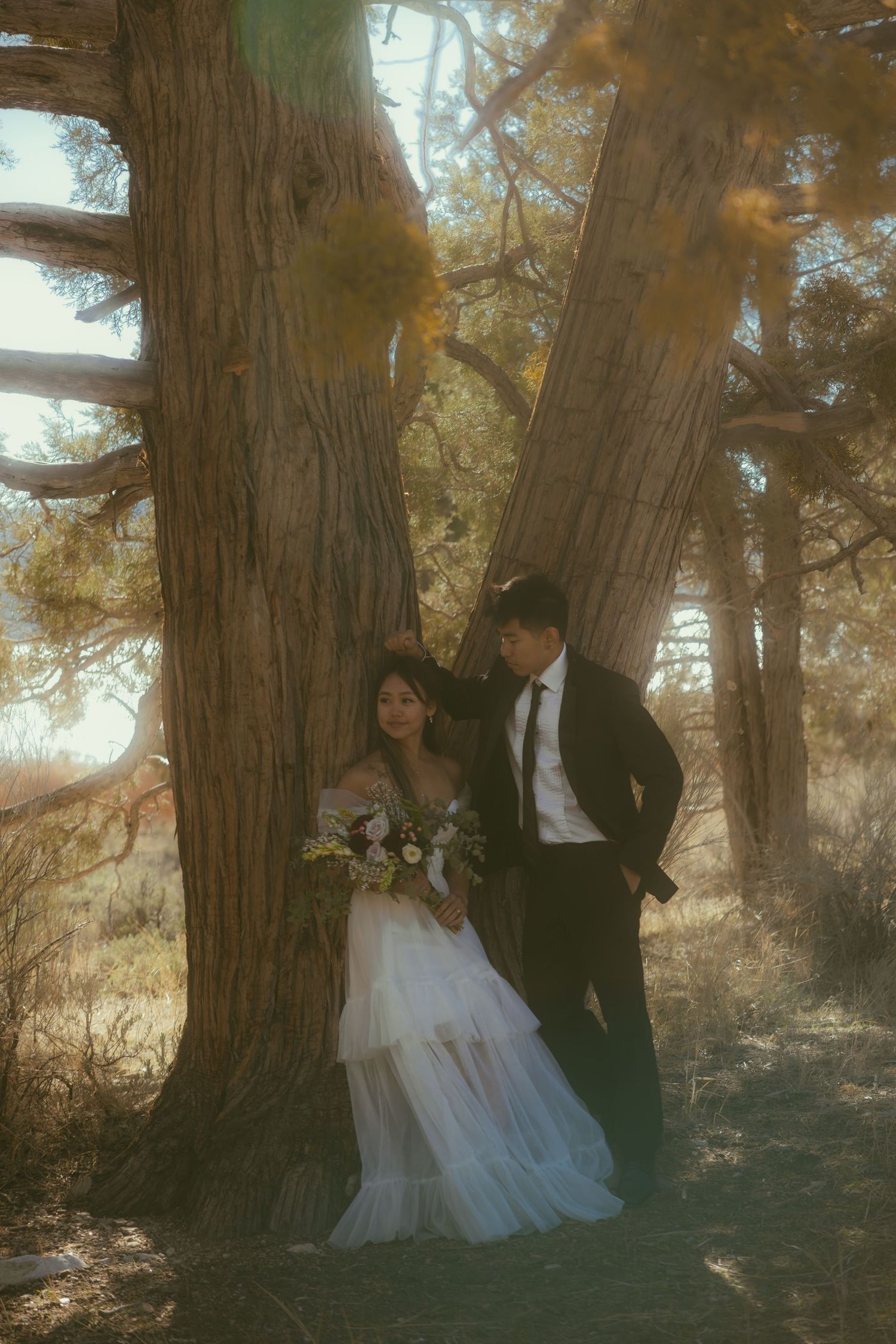 Bride and groom pose romantically against a large tree trunk in a sunlit forest, bride holding a white floral bouquet.