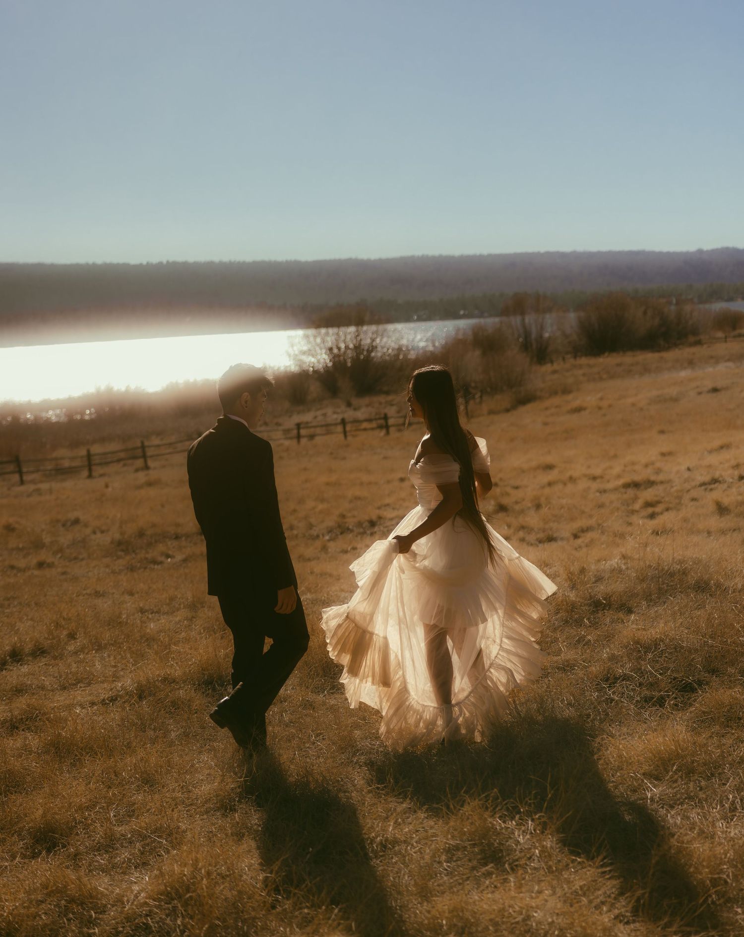 Couple walking through golden field at sunset near a lake, bride in flowing tulle gown, groom in dark suit.
