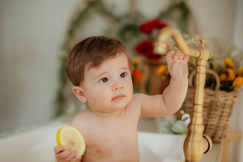 Cute baby sitting in a white bathtub holding a lemon slice, reaching up to touch a gold faucet.