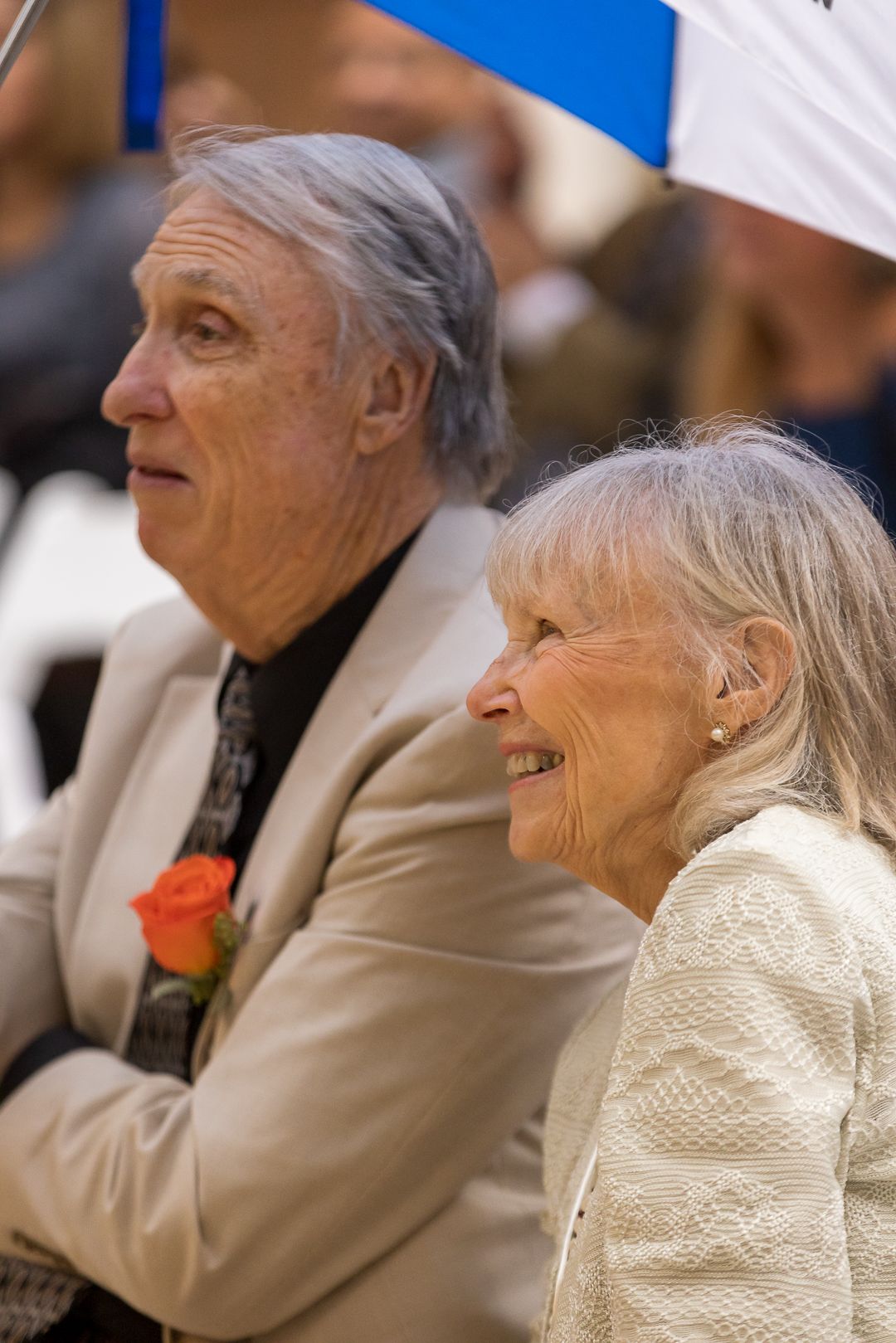 Elderly couple smiling at an outdoor event, man in white blazer with orange boutonniere, woman in cream jacket.