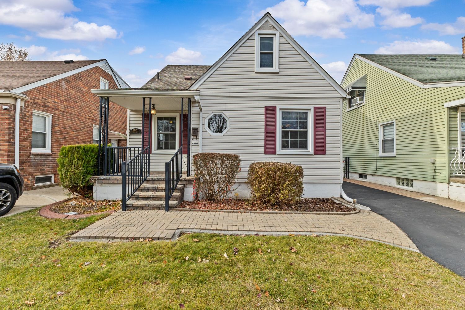 Cape cod style home with gray siding burgundy shutters covered front porch and paved driveway.