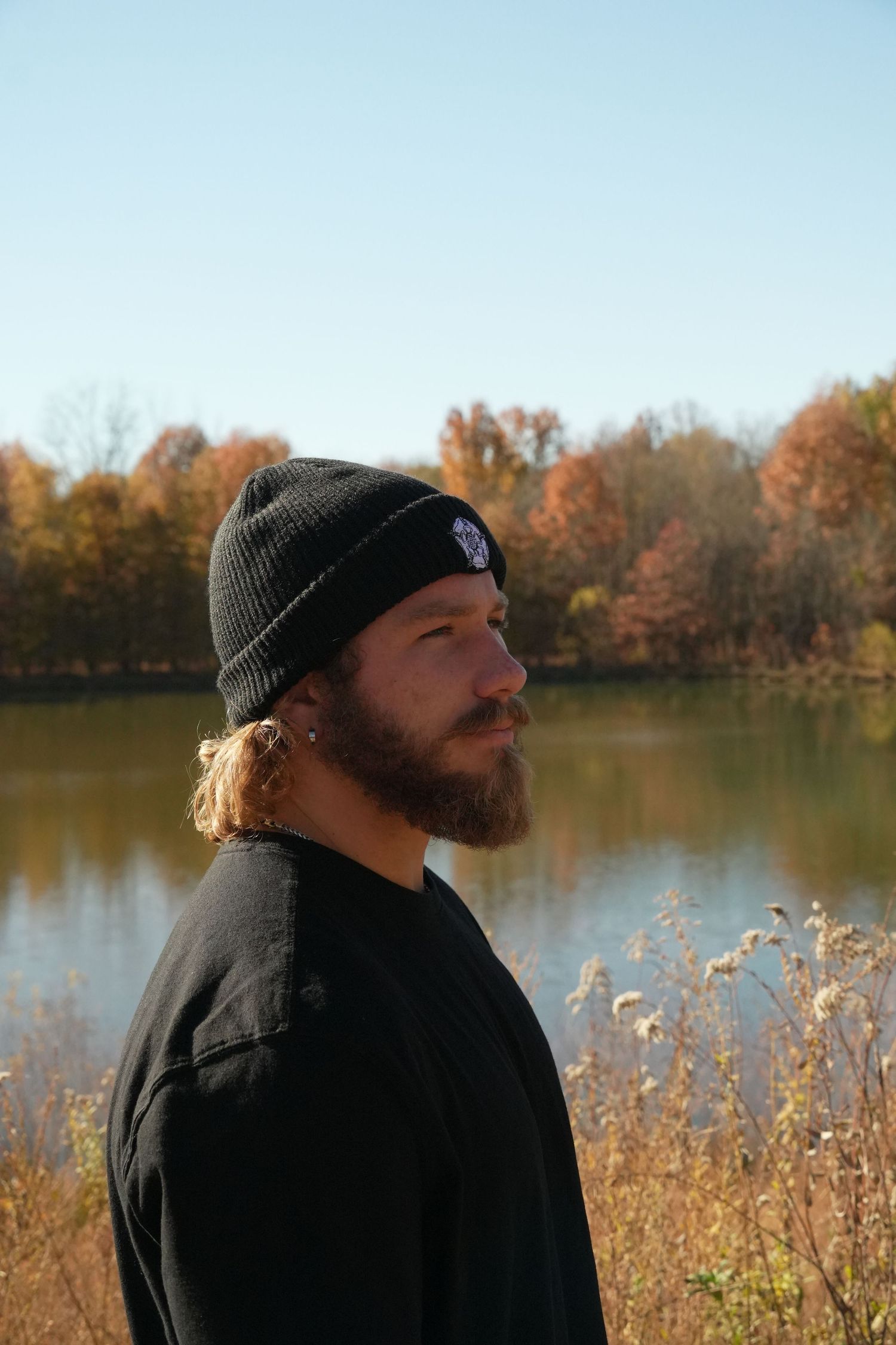 Bearded man in black beanie and jacket standing by a calm lake surrounded by autumn foliage on a clear day.