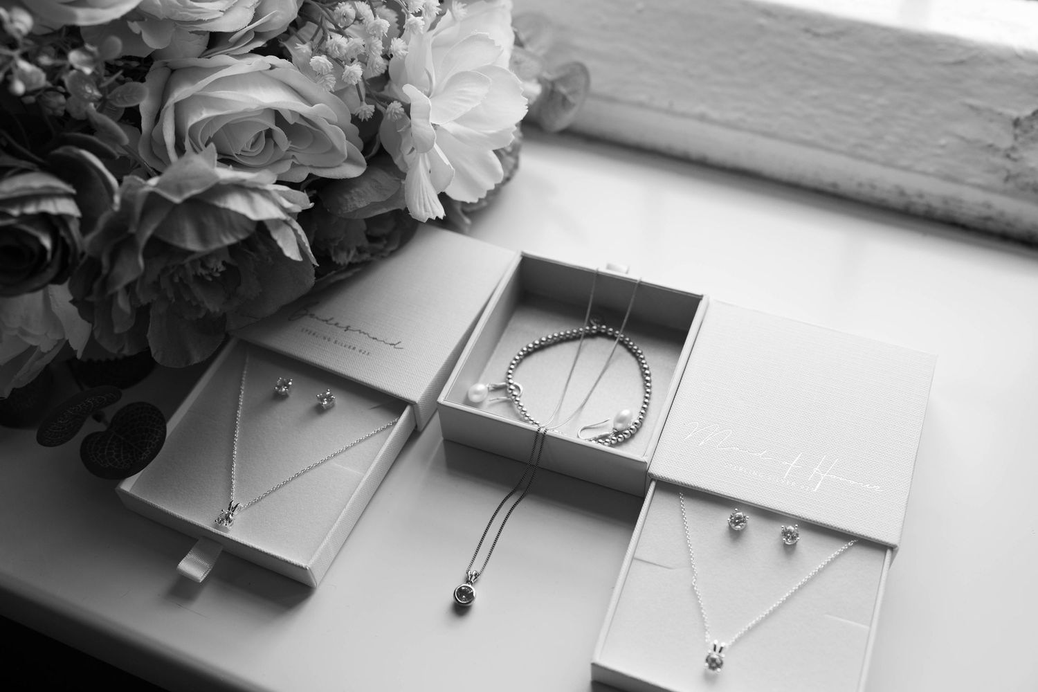 Black and white flat lay of elegant bridal jewelry sets in gift boxes beside floral bouquet.