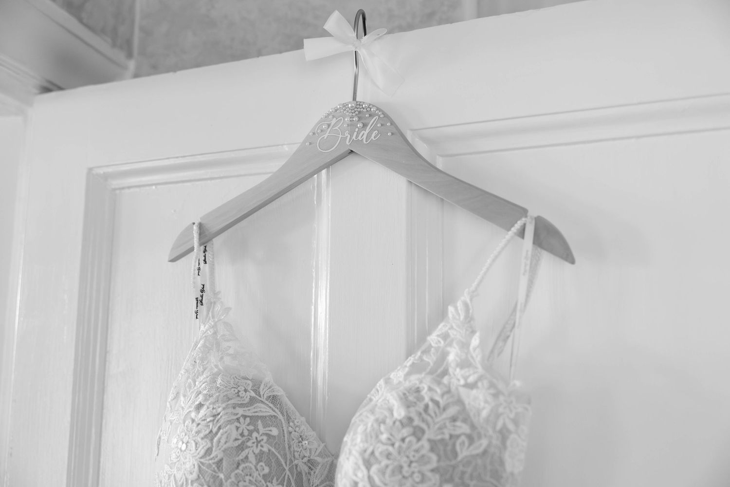 Delicate lace bridal bralette hanging on decorative wooden hanger against white door in black and white.