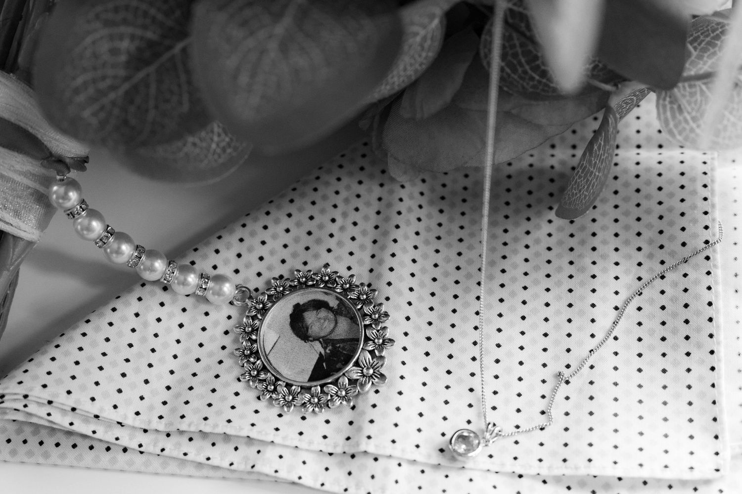 Black and white close-up of a memorial photo locket charm beside pearl bracelet on polka dot fabric.