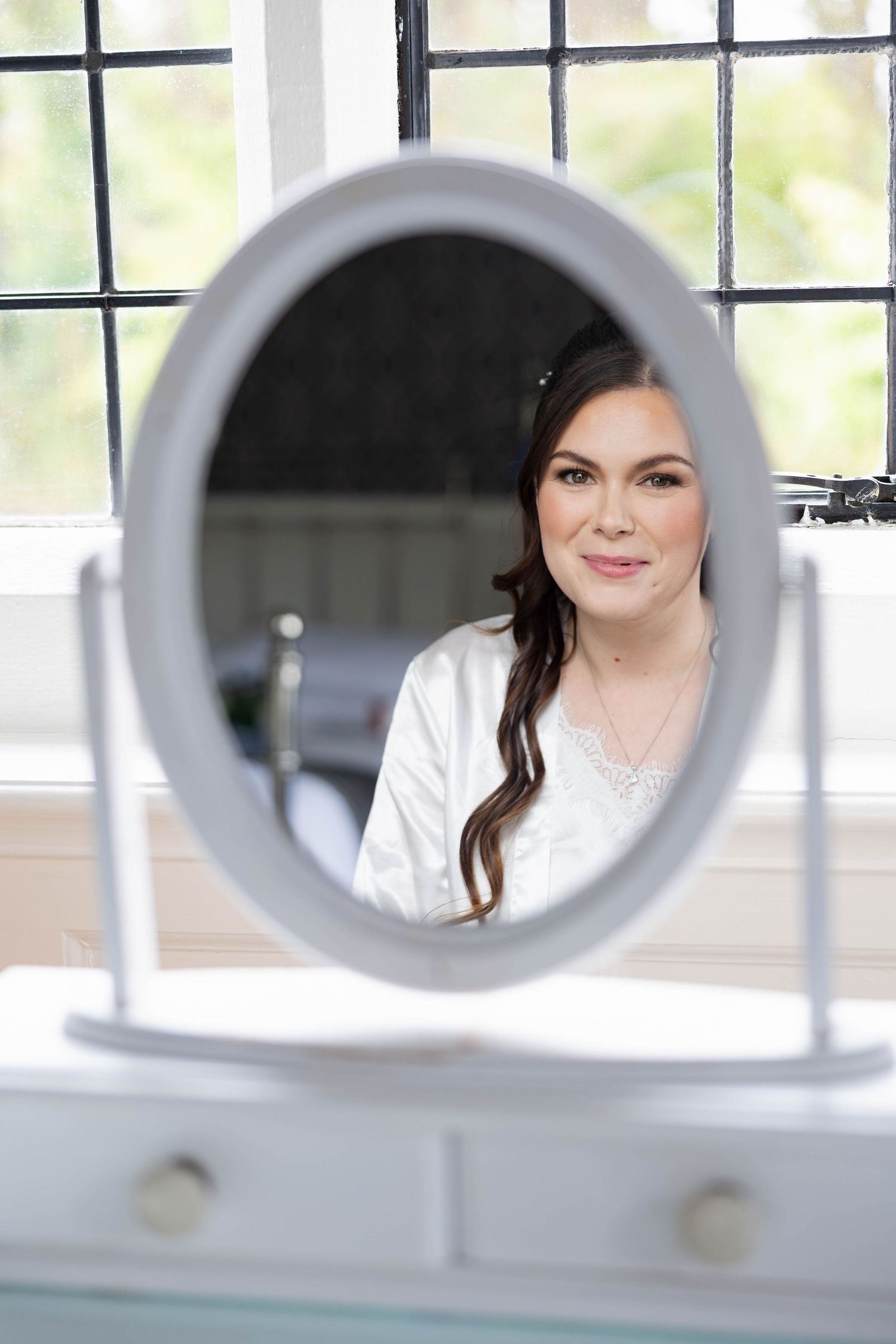 Beautiful bride in white robe smiles softly at her reflection in oval vanity mirror before wedding.