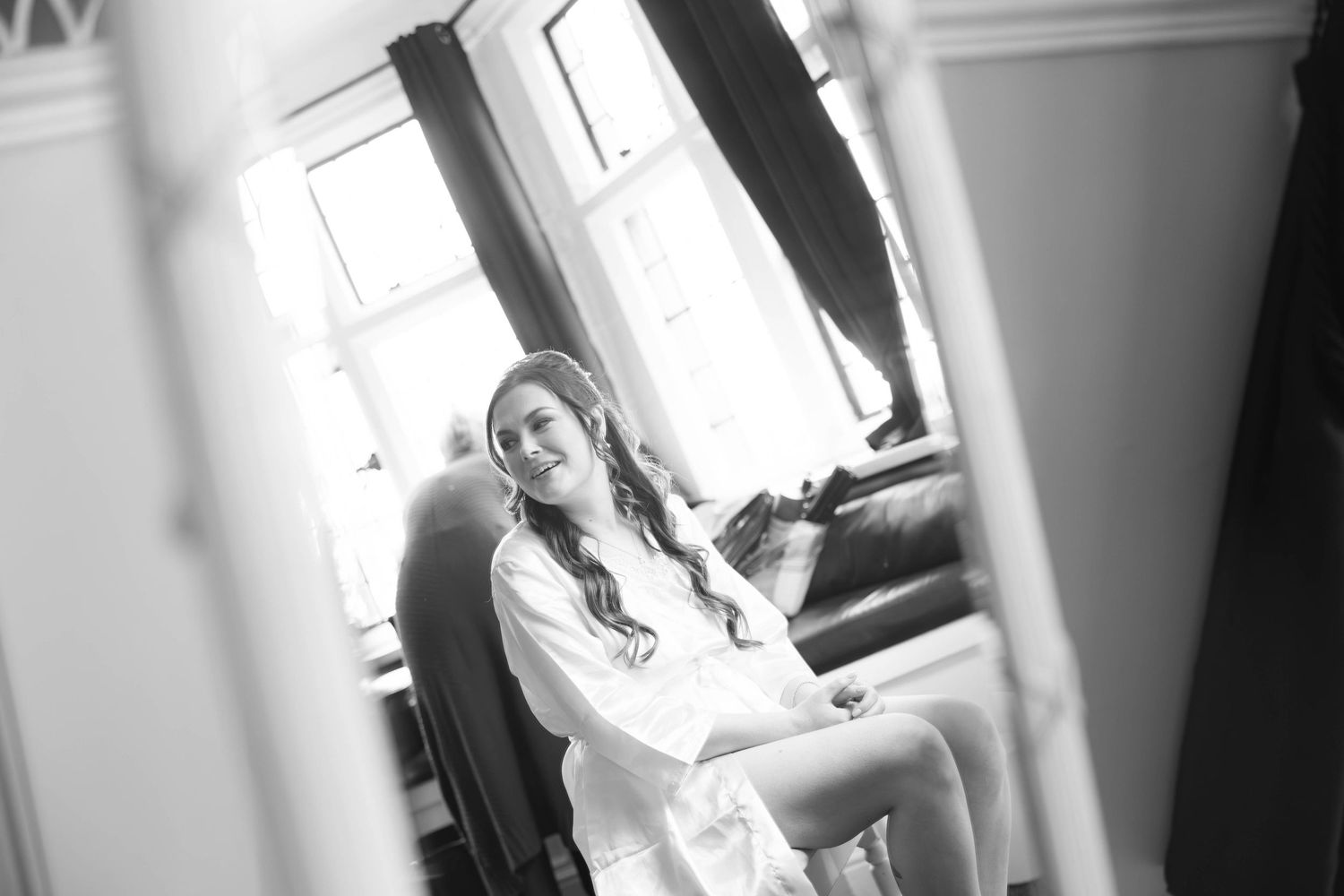 Black and white photo of smiling bride in white robe relaxing on sofa during wedding morning preparations.