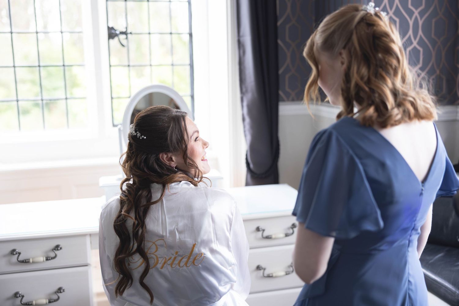 Bride in white robe chatting with bridesmaid in blue dress by sunlit window during wedding morning preparations.
