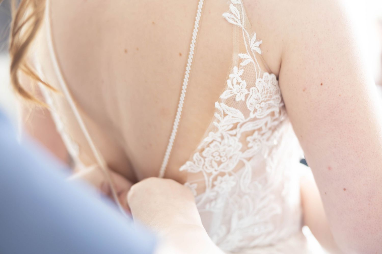 Close-up detail of lace wedding dress back being fastened with pearl button closure by bridesmaid.