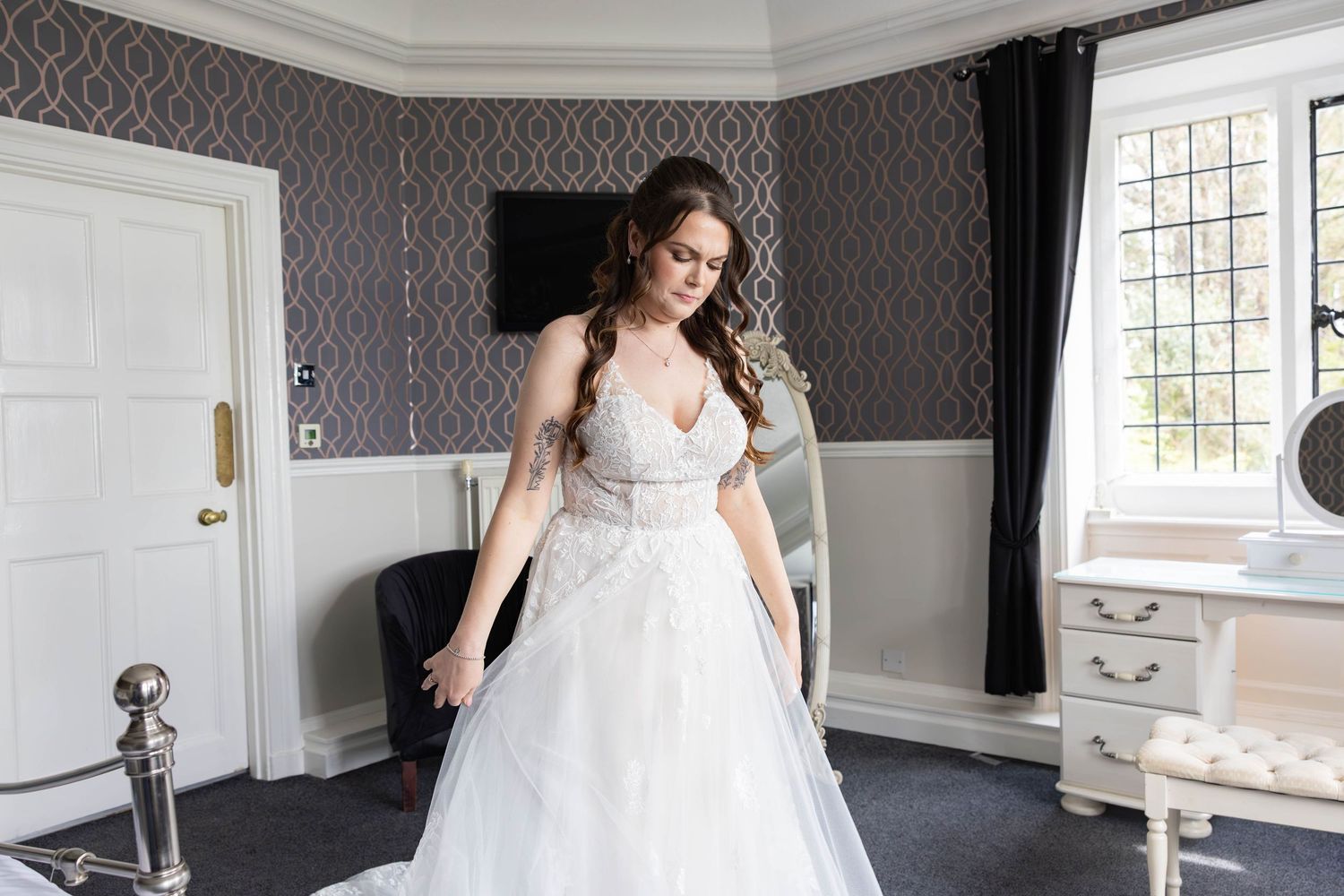 Bride in elegant lace and tulle ballgown wedding dress standing in ornate grey decorated bridal suite.