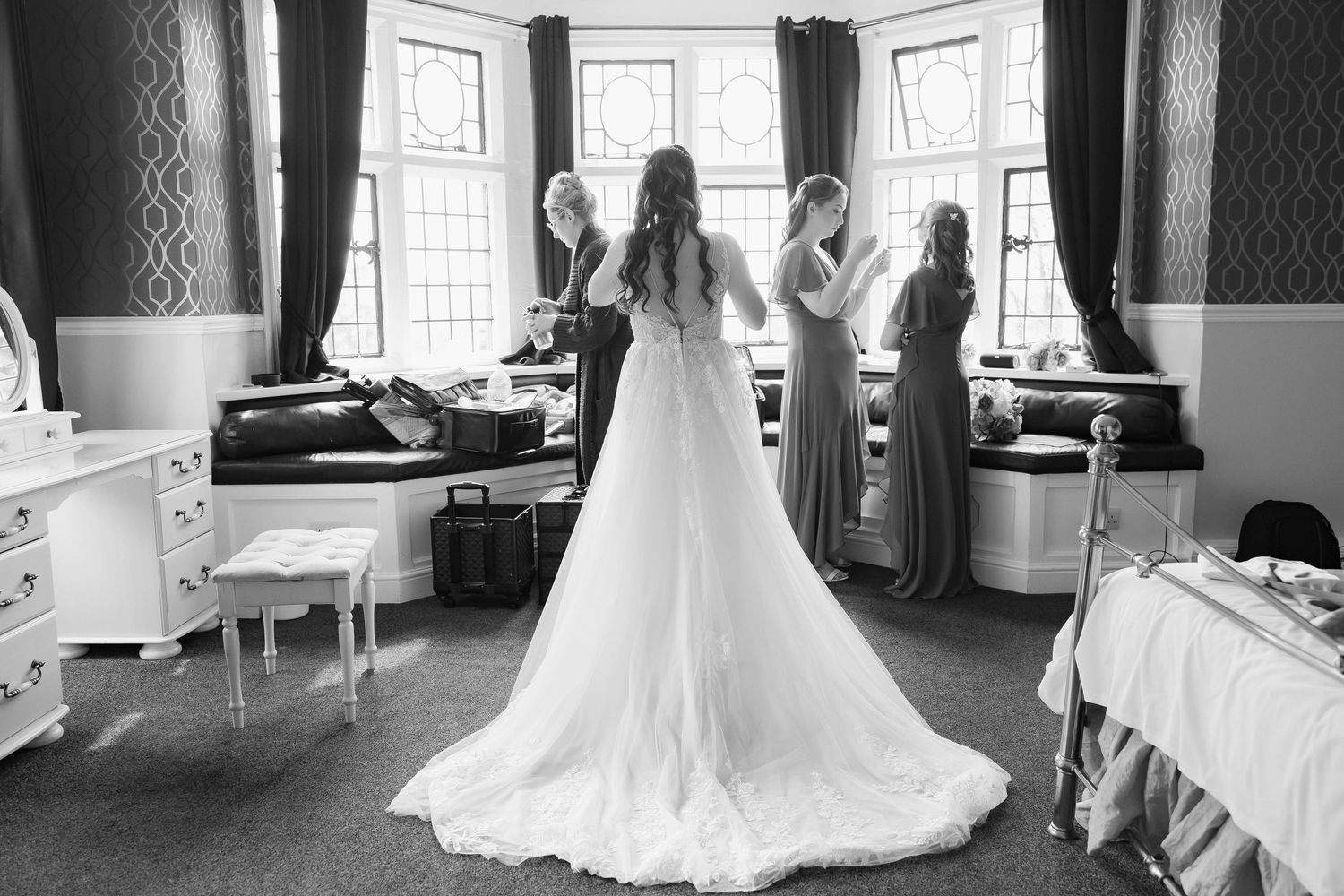 Black and white photo of bride in full tulle gown with bridesmaids holding up her train by window.