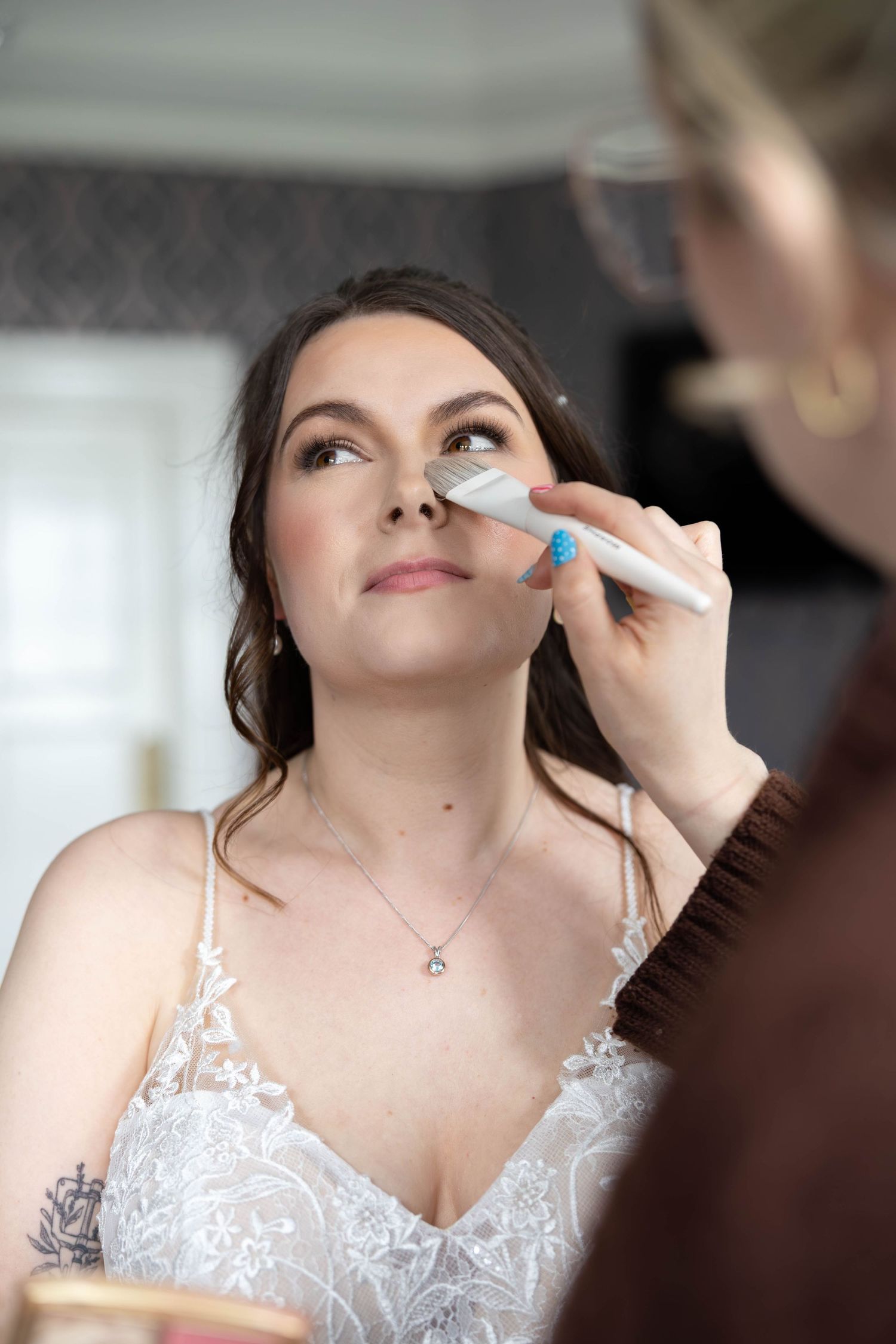 Makeup artist applying foundation to a bride in a lace wedding dress before her wedding ceremony.