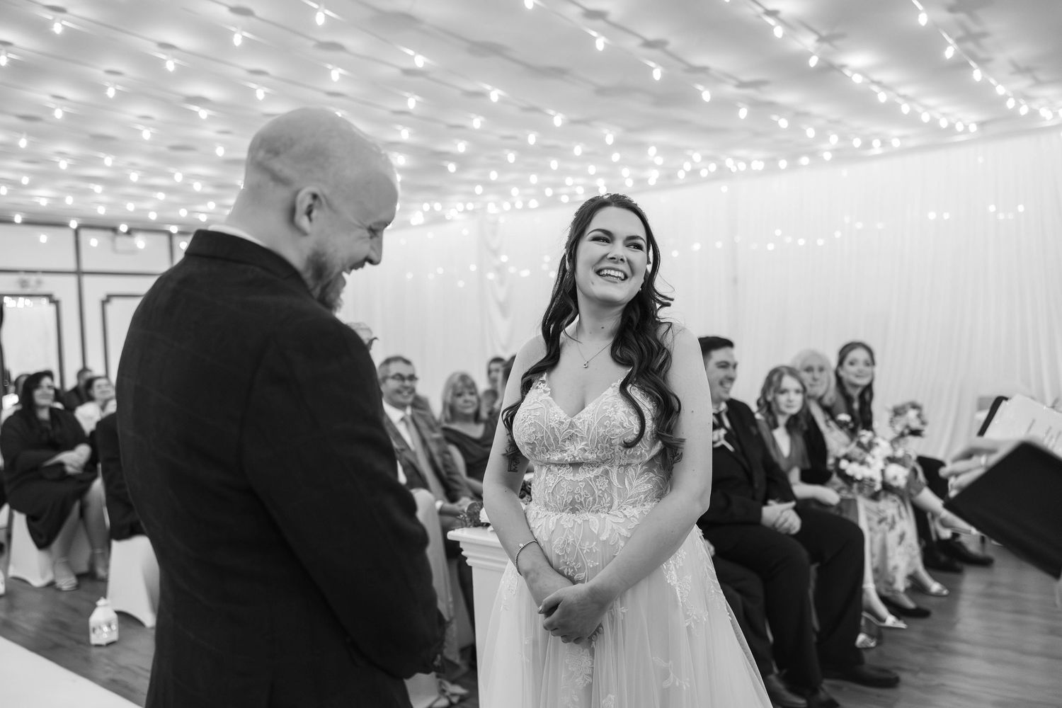 Smiling bride in a beaded gown exchanging vows with groom under fairy lights at wedding ceremony.