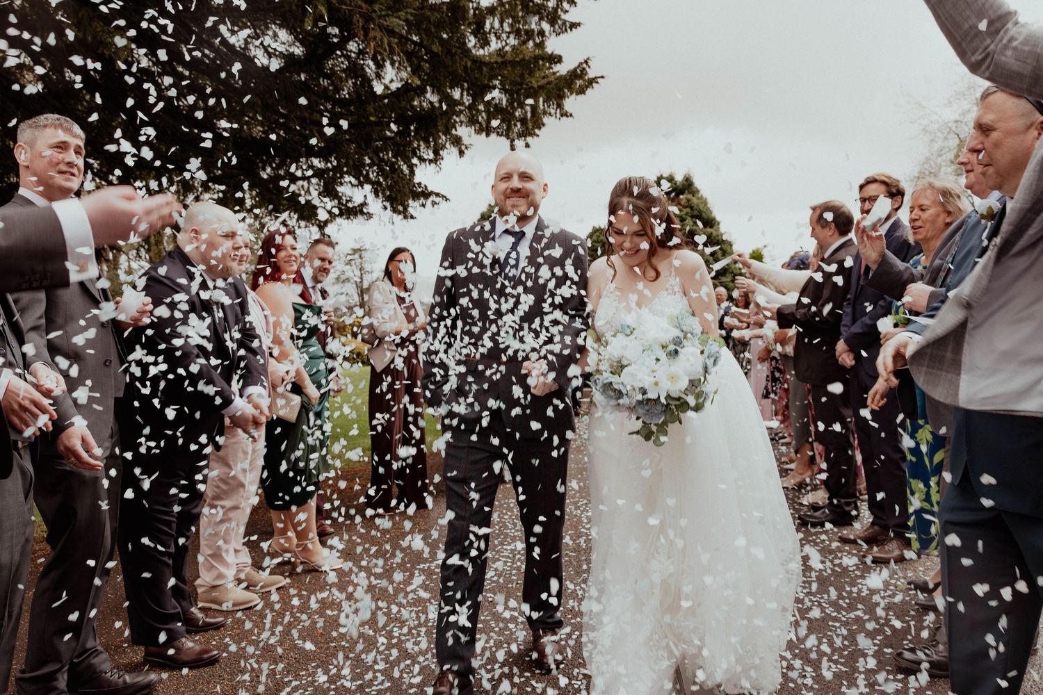 Bride and groom walking through a colorful confetti shower surrounded by cheering wedding guests.