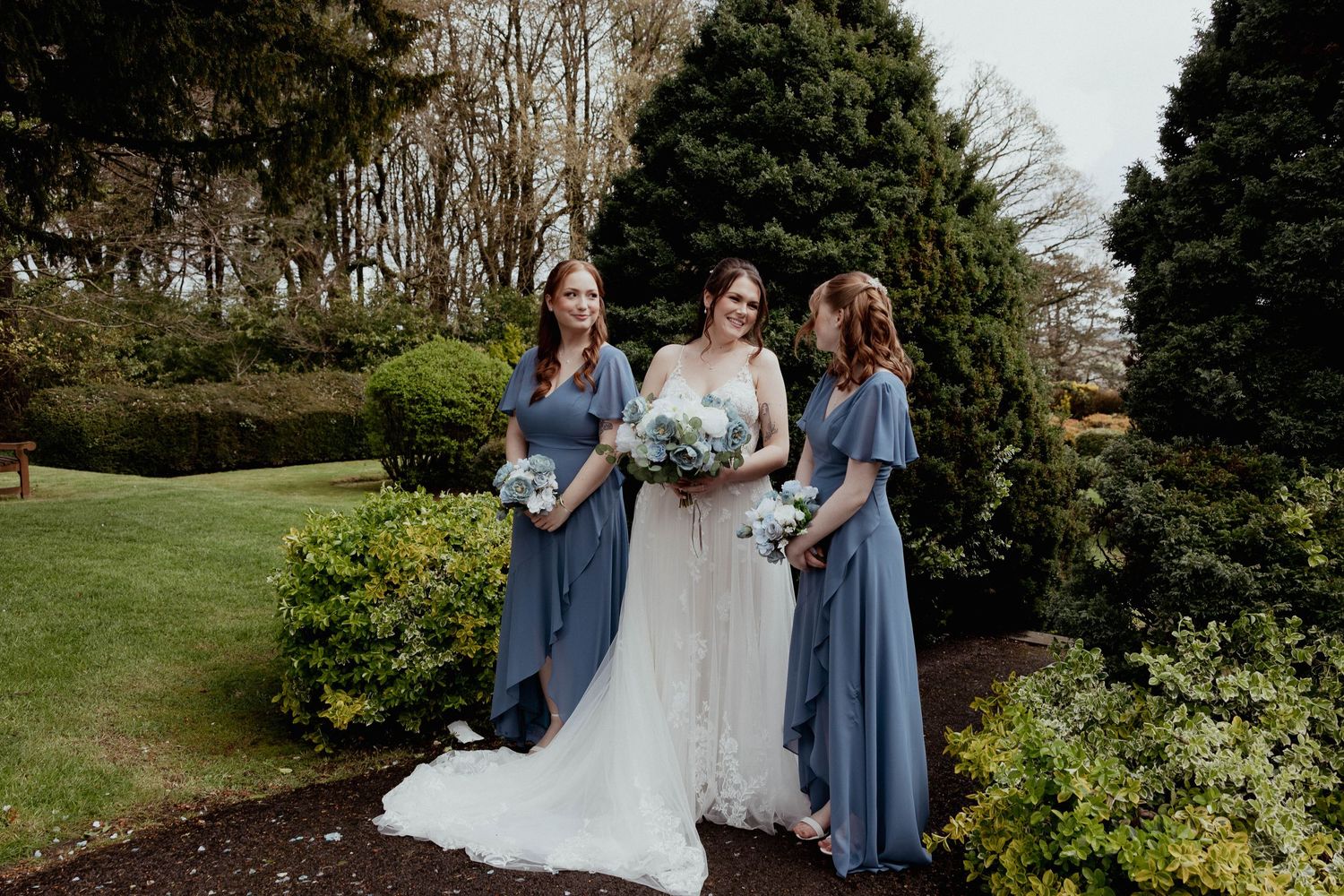 Bride in lace wedding gown posing with two bridesmaids in dusty blue dresses in a garden.