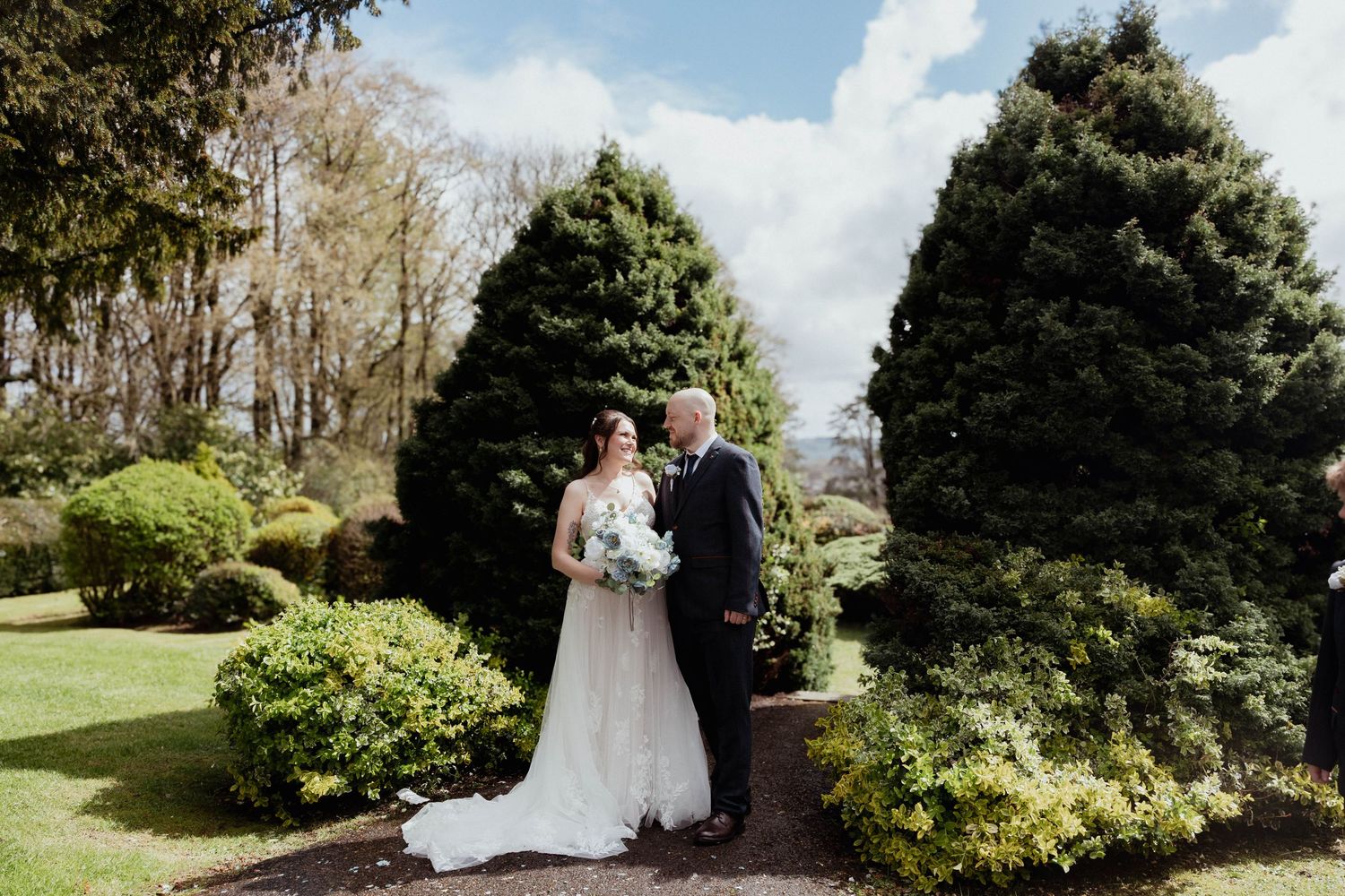 Bride and groom sharing a romantic moment on a garden path surrounded by tall evergreen trees.