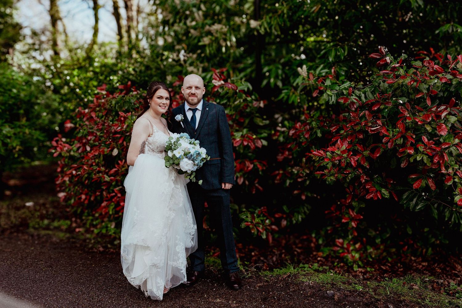 Bride and groom posing together outdoors in front of lush red and green foliage.