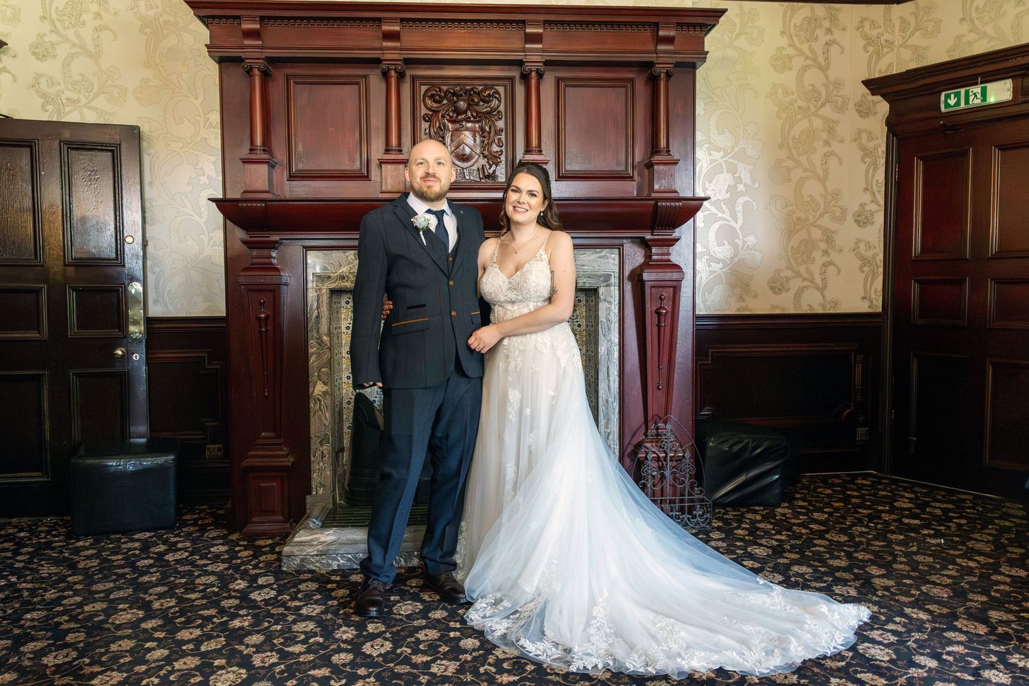 Bride and groom posing elegantly in front of a ornate wooden fireplace in a grand venue.