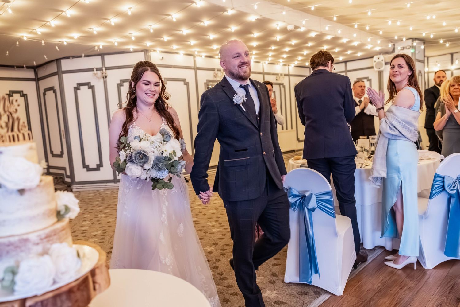 Newlywed couple walking hand in hand into their wedding reception under warm fairy lights.