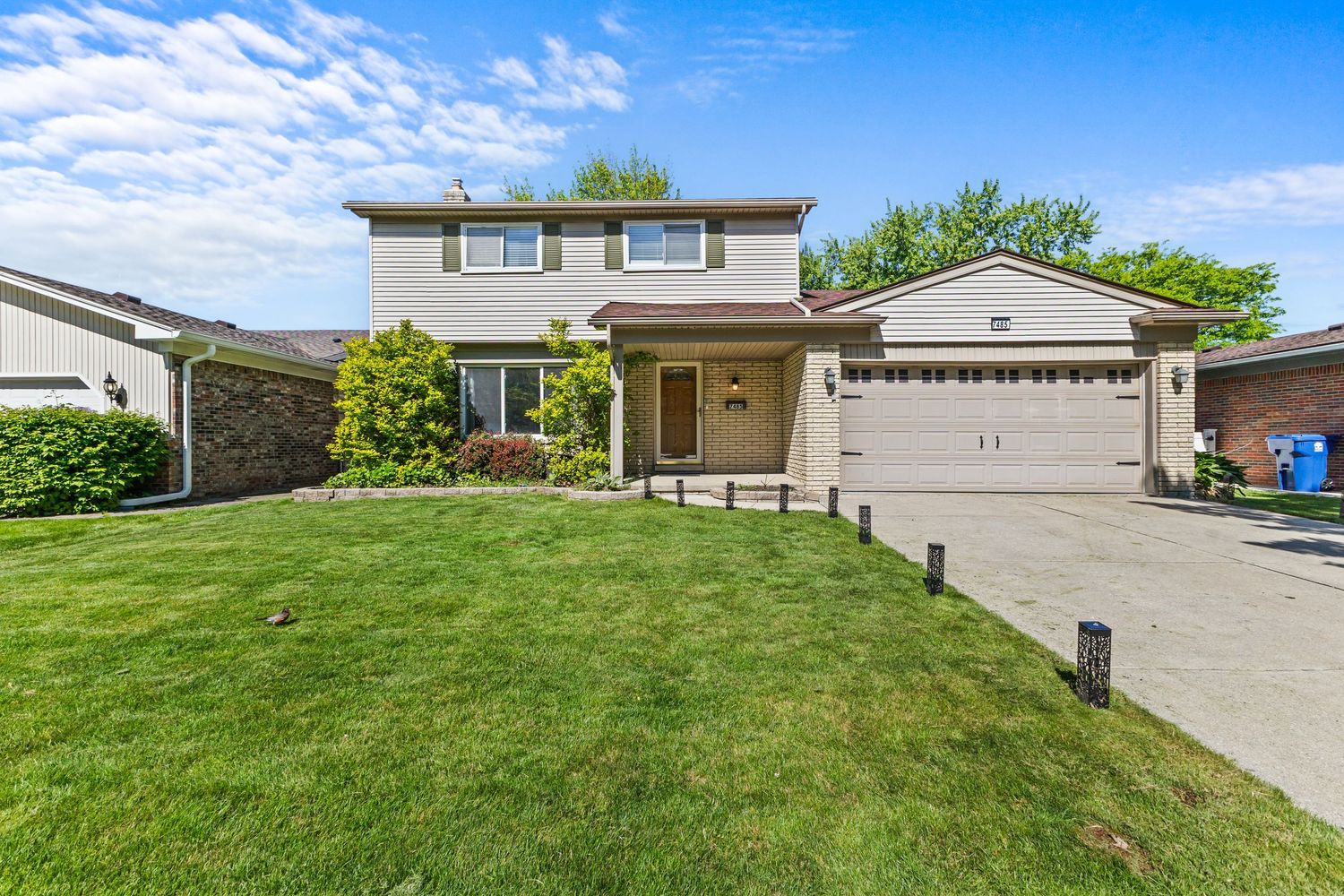 Two-story beige home with attached two-car garage, landscaped front yard, and clear blue sky.