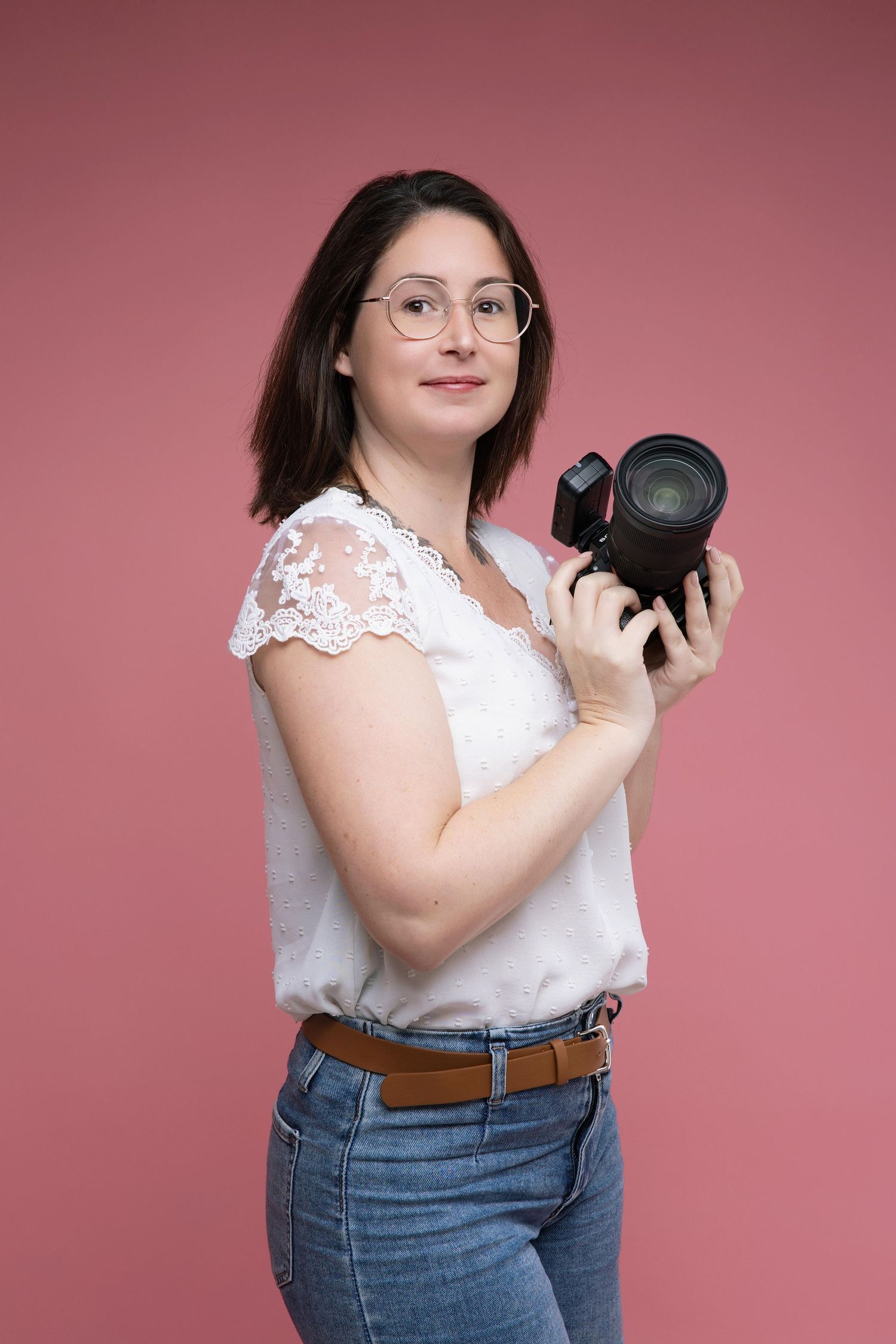 Professional photographer wearing glasses and lace top holds DSLR camera against pink studio backdrop.