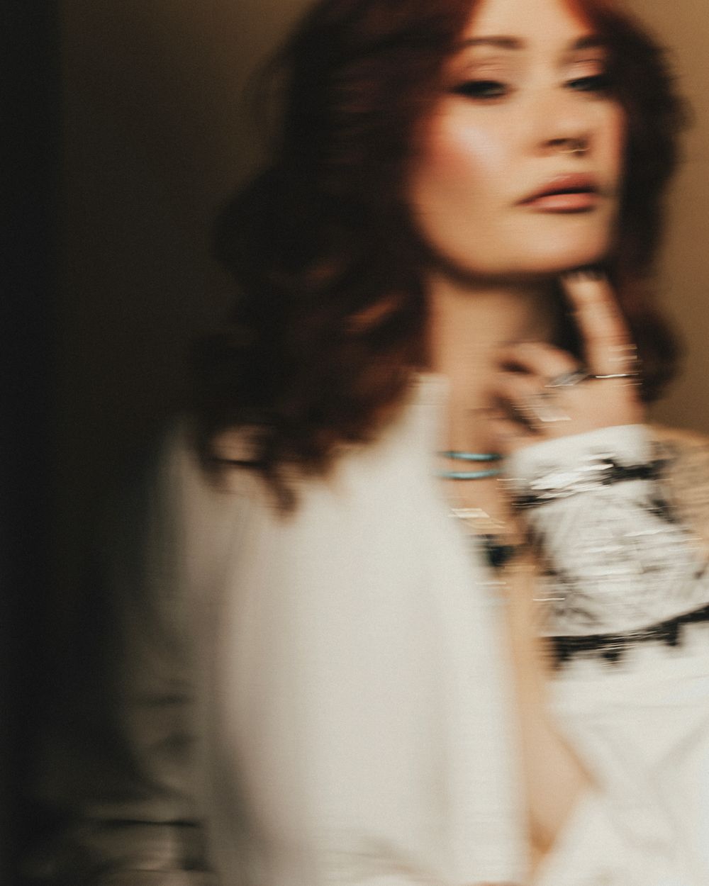 Close-up portrait of a redhead woman wearing silver jewelry and white outfit in a softly blurred editorial photo.