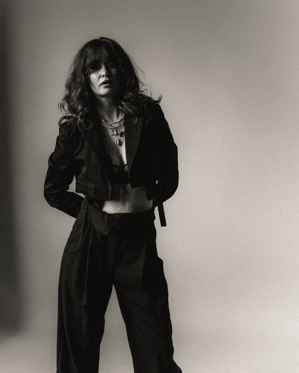 Black and white photo of a woman in a cropped jacket and wide-leg trousers, posing confidently with layered necklaces.