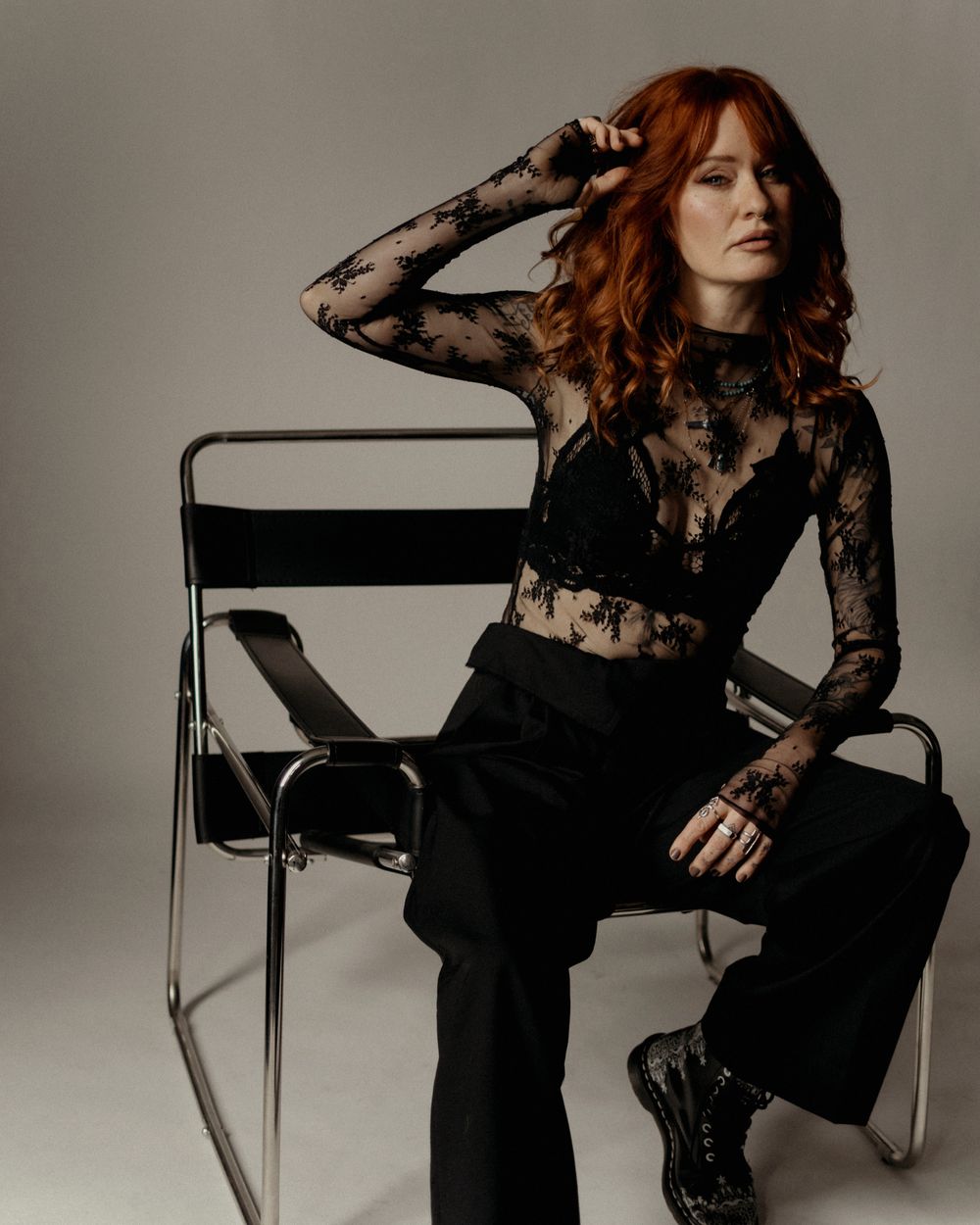 Woman with red hair poses in black lace top and trousers on a chrome chair.