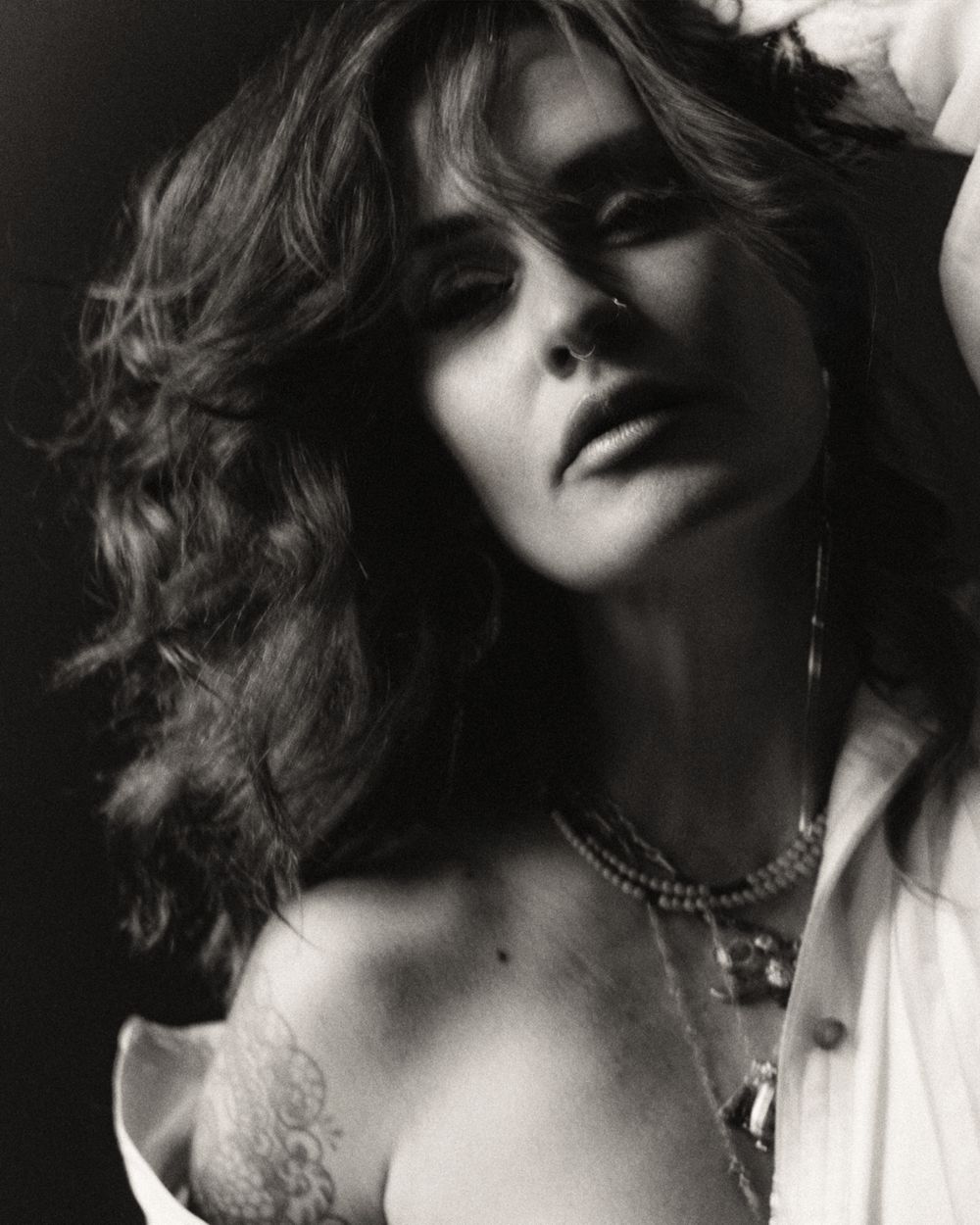Black and white portrait of a woman with wavy hair, wearing layered necklaces and an open white shirt.