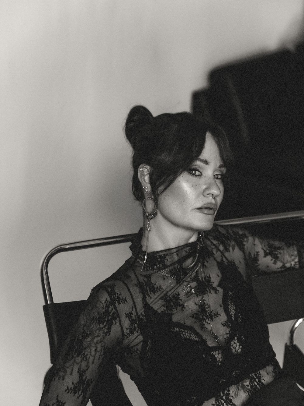 Black and white photo of a woman in lace top with bun hairstyle and hoop earrings, seated in a chair.