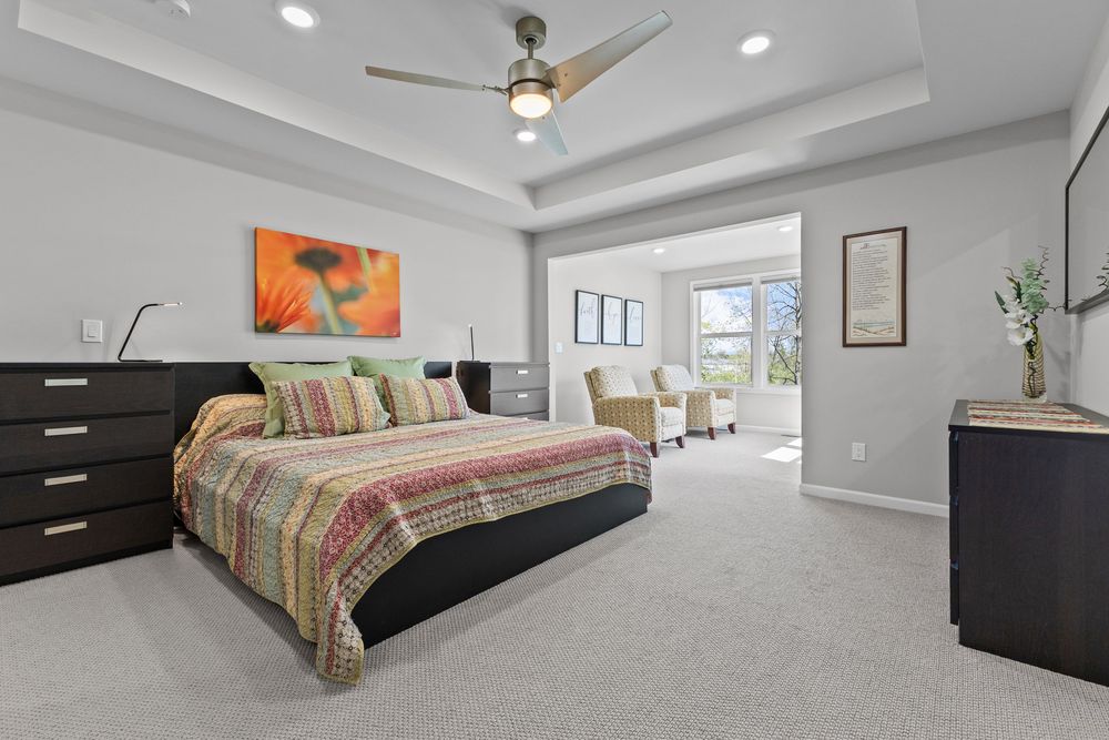 Elegant master bedroom with tray ceiling, plush carpet, ceiling fan, and sitting area with natural light.