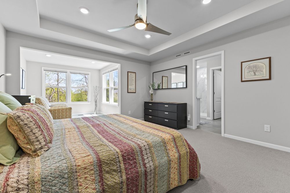 Spacious master bedroom with tray ceiling, ceiling fan, plush carpet, colorful quilt, and bright windows leading to sitting area.