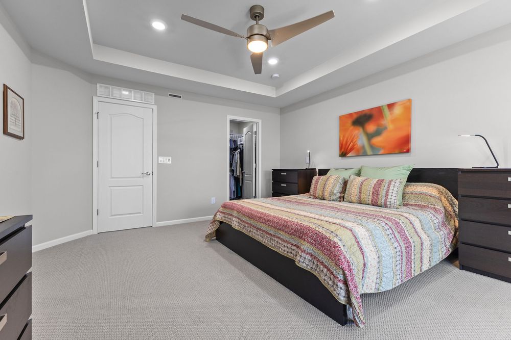 Elegant master bedroom featuring tray ceiling, ceiling fan, dark wood dresser, and vibrant floral artwork above the bed.