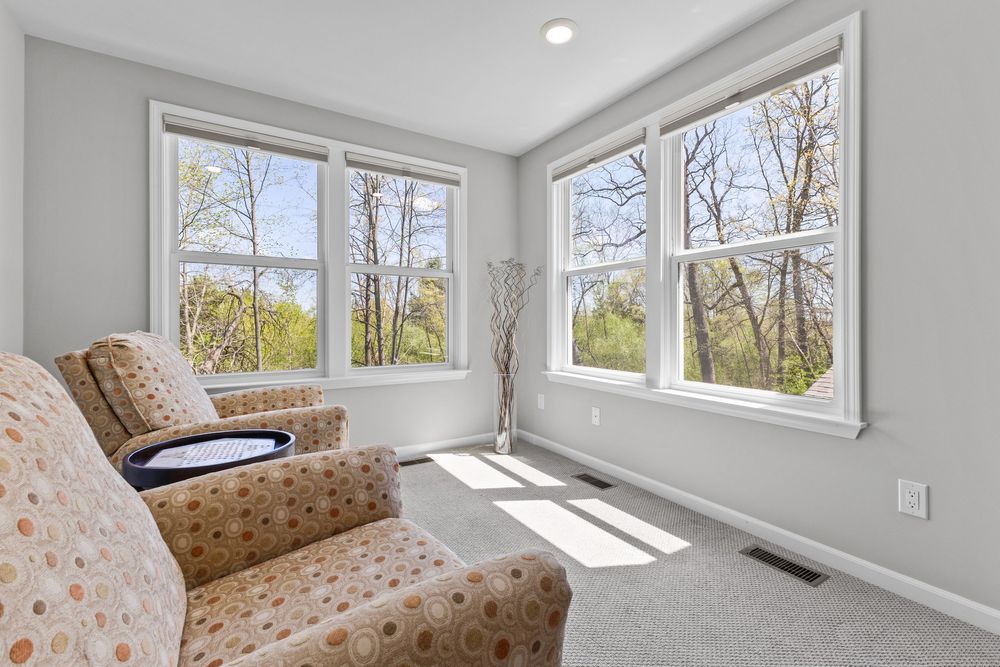 Bright sunroom sitting area with large windows overlooking wooded backyard, featuring two patterned armchairs and carpet flooring.