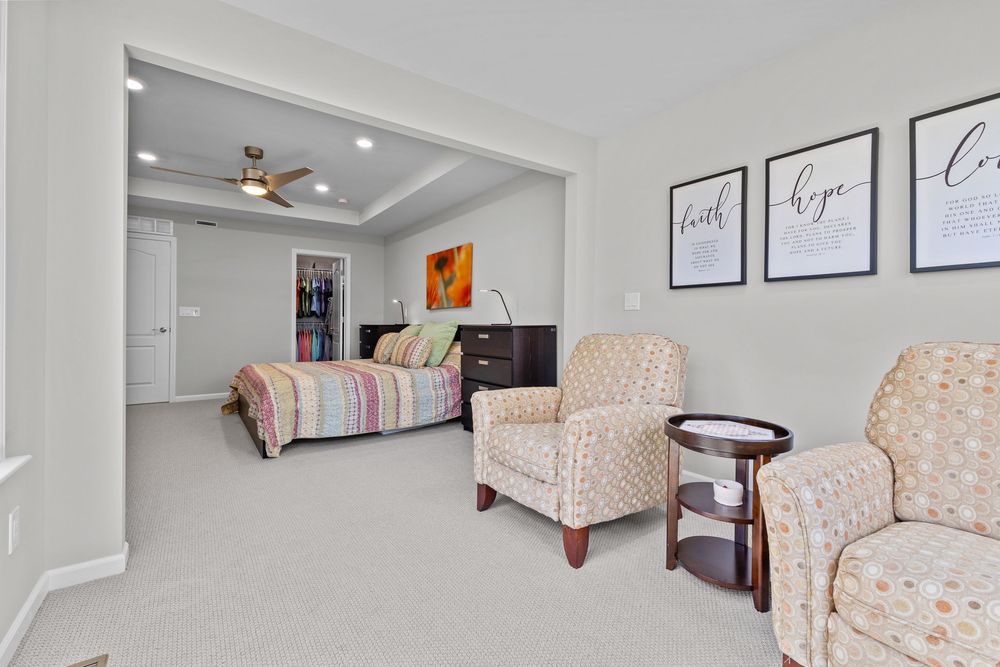 Open bedroom suite with tray ceiling, ceiling fan, patterned armchairs, and colorful striped bedding on a dark wood bed.