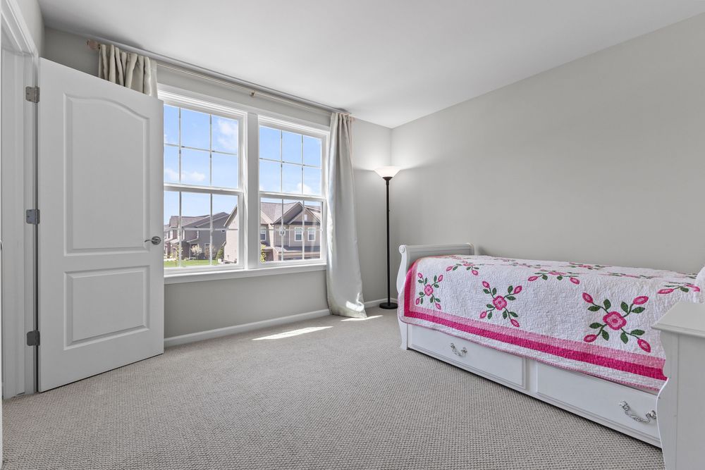 Cozy bedroom with white daybed featuring floral quilt, large window with neighborhood views, and light gray carpet flooring.