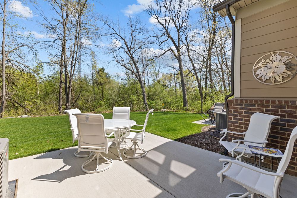 Outdoor patio with white dining table and chairs, gas grill, and serene wooded backyard with lush green lawn.